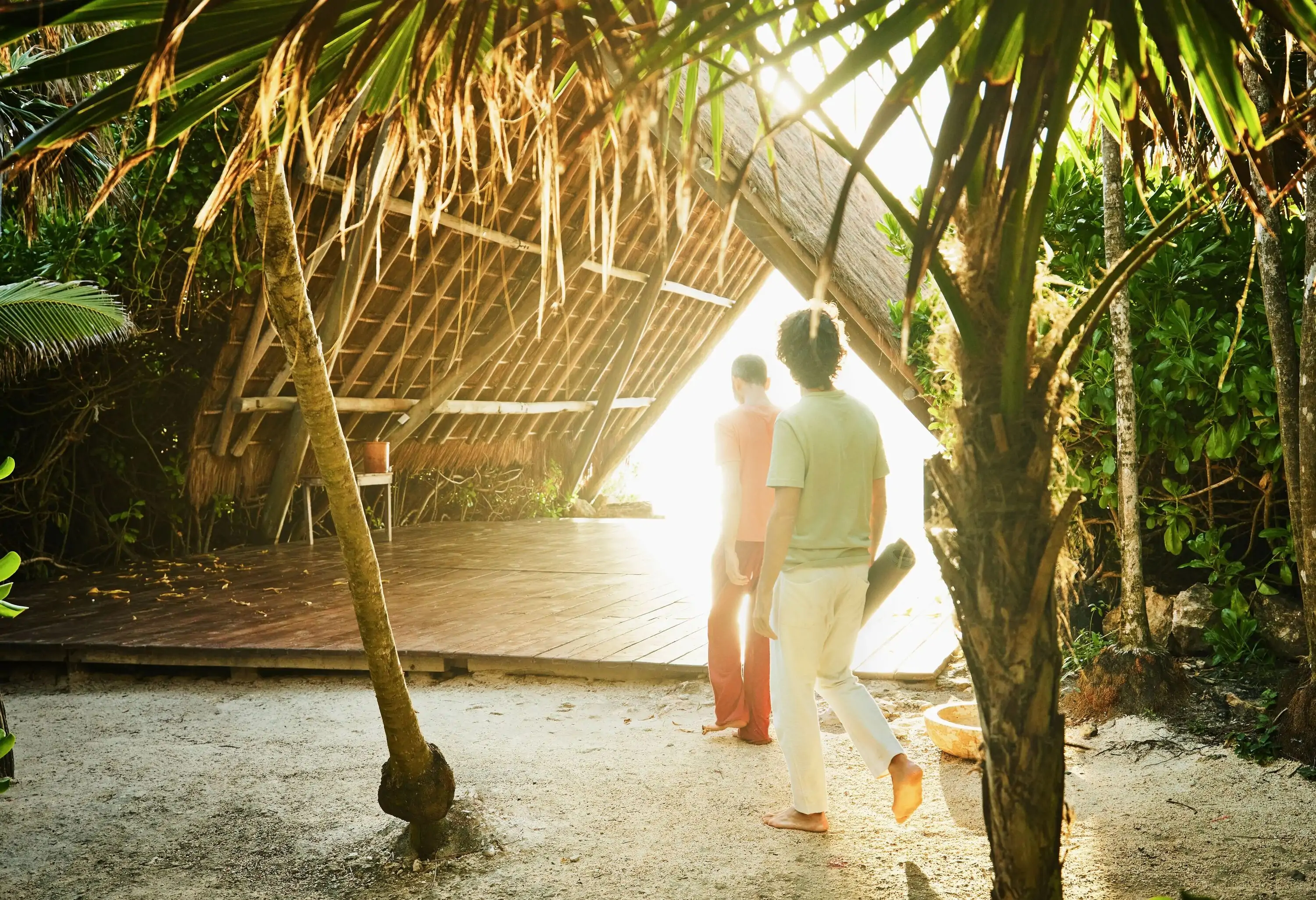 Two people walk through a jungle towards a wooden covered platform while carrying a yoga mat.