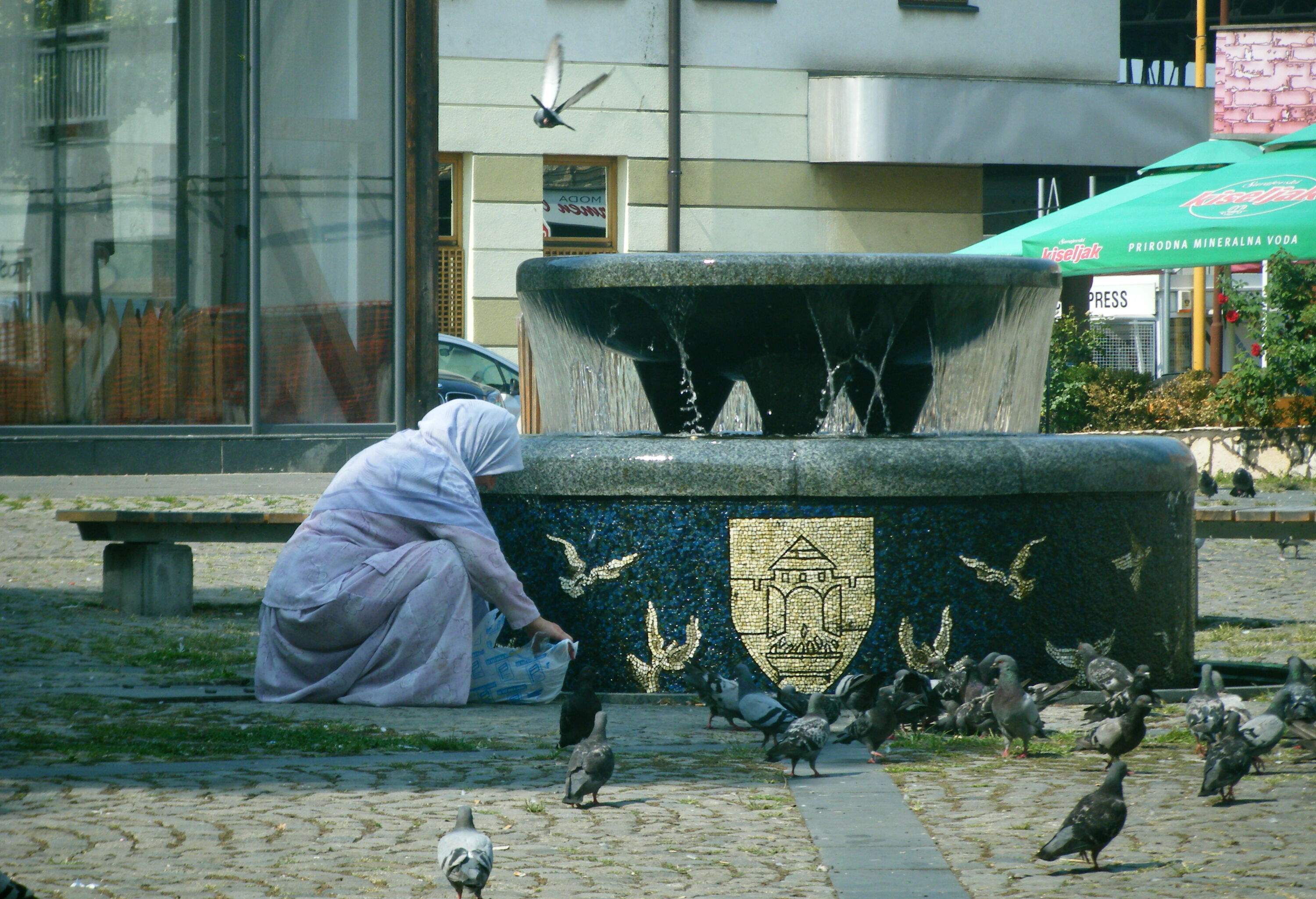 A woman feeding the pigeons beside a fountain with food from a plastic bag.