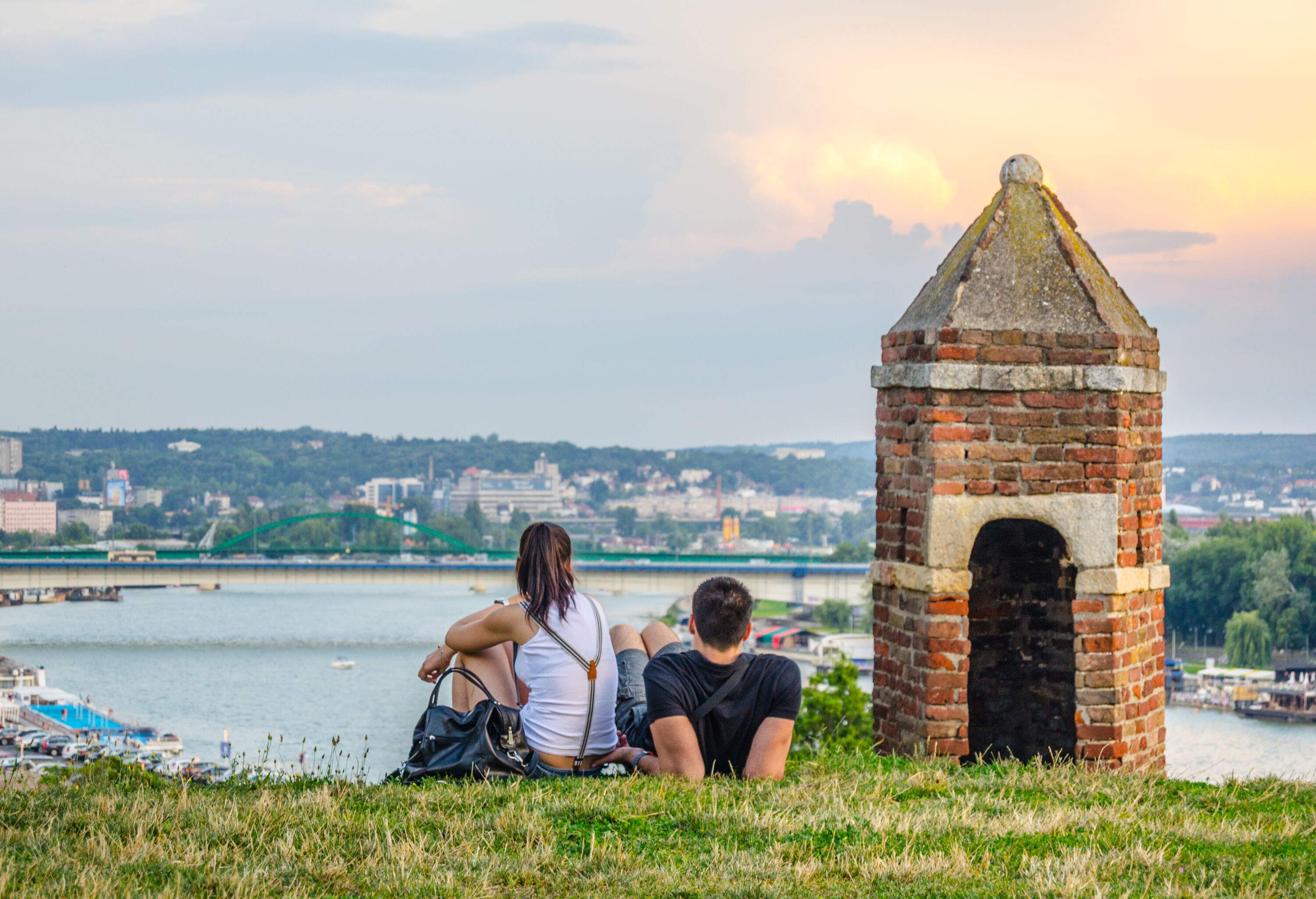 A man and a woman lounging next to a brick tower on a grassy hill overlooking a river.