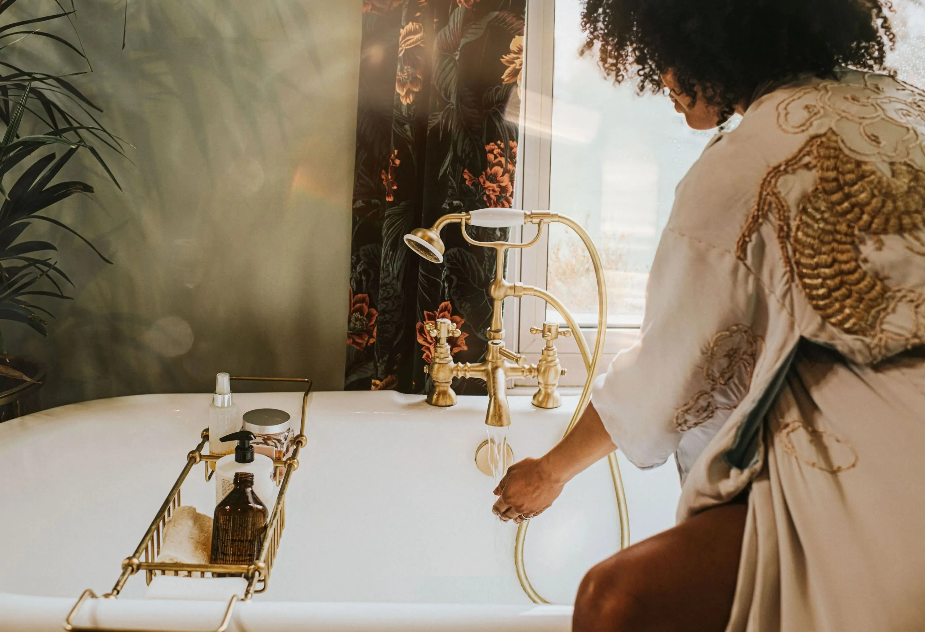 A beautiful young woman holds her hands under the gold faucet of a roll top bath to access the temperature of the water as she fills the tub. The scene is relaxed, and luxurious.