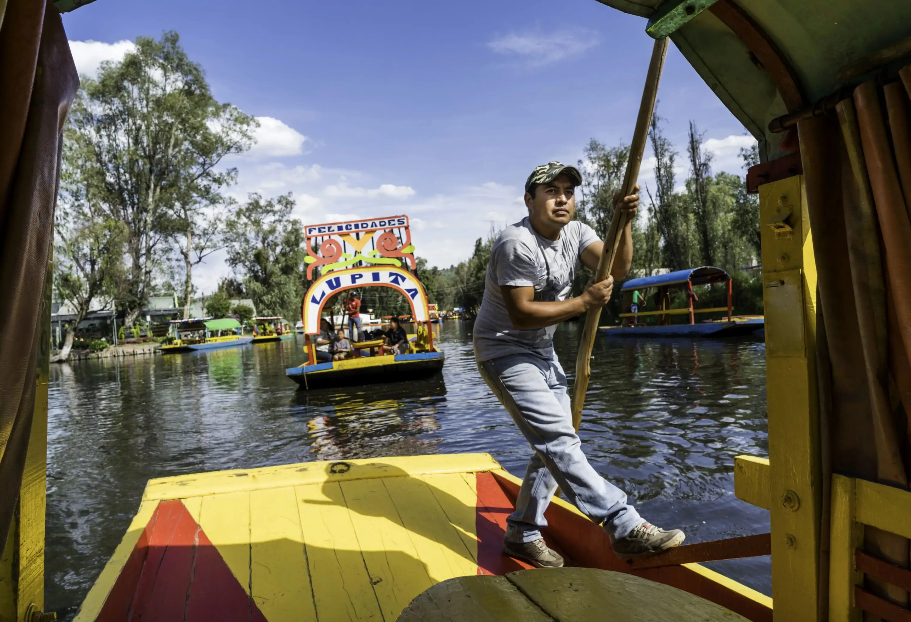 A man holding on to a wooden post on a traditional Mexican trajinera as it cruises along the canal.