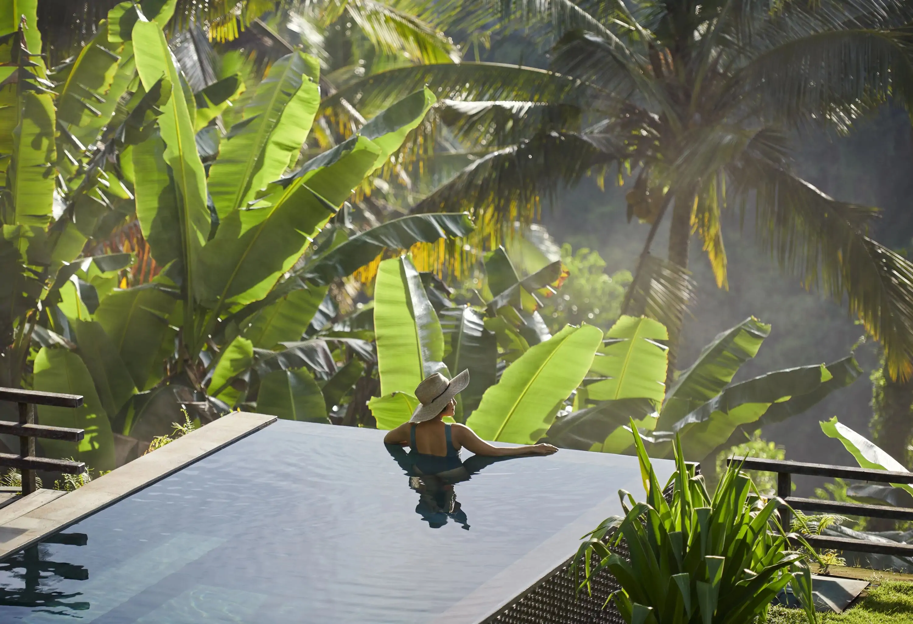 Asian woman enjoying beautiful private pool at luxury villa in Ubud, surrounded by tropical trees and plants, situated on edge of misty valley