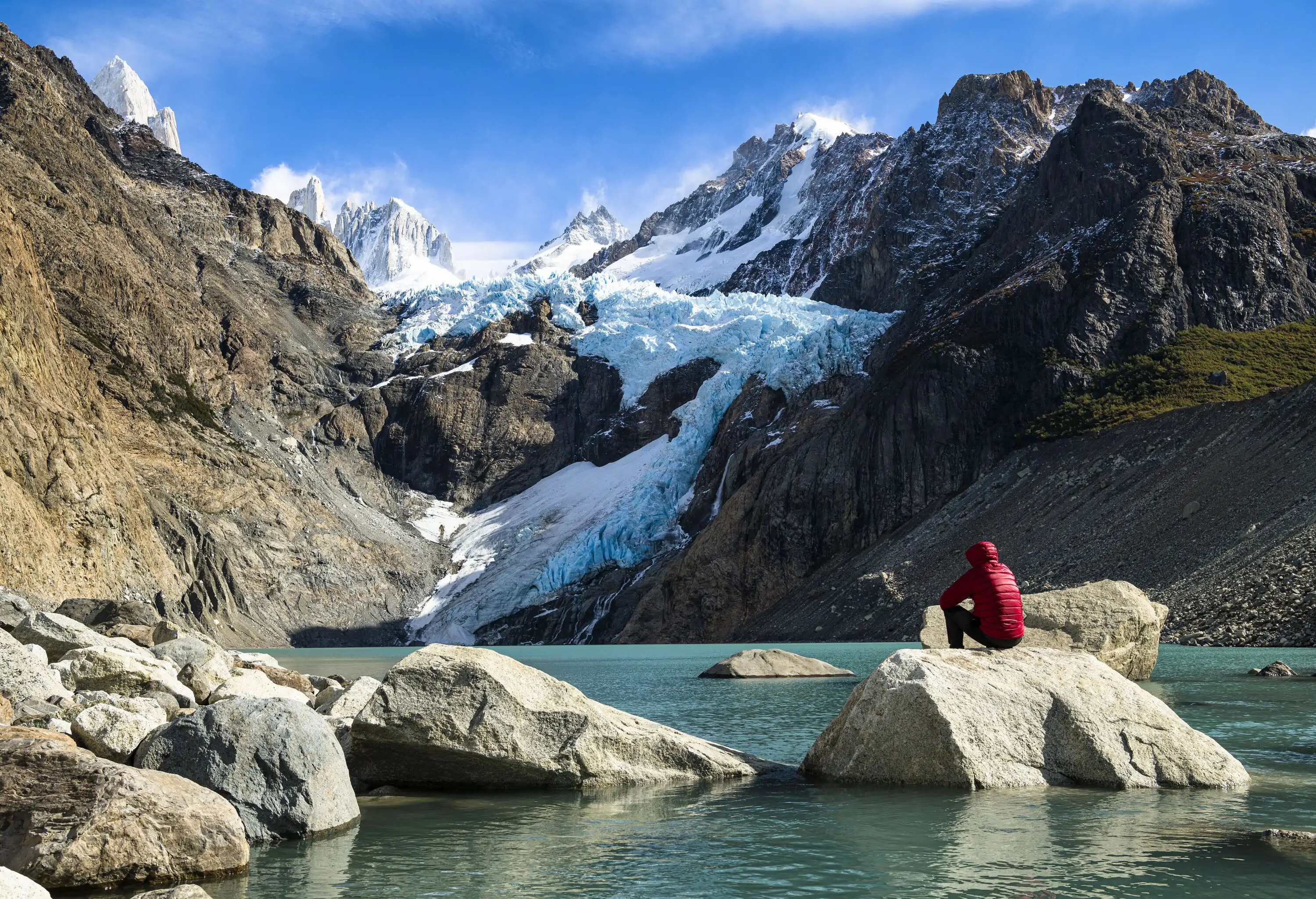 A man in a red jacket sitting on a huge rock in a lake, staring out towards snowy mountains.