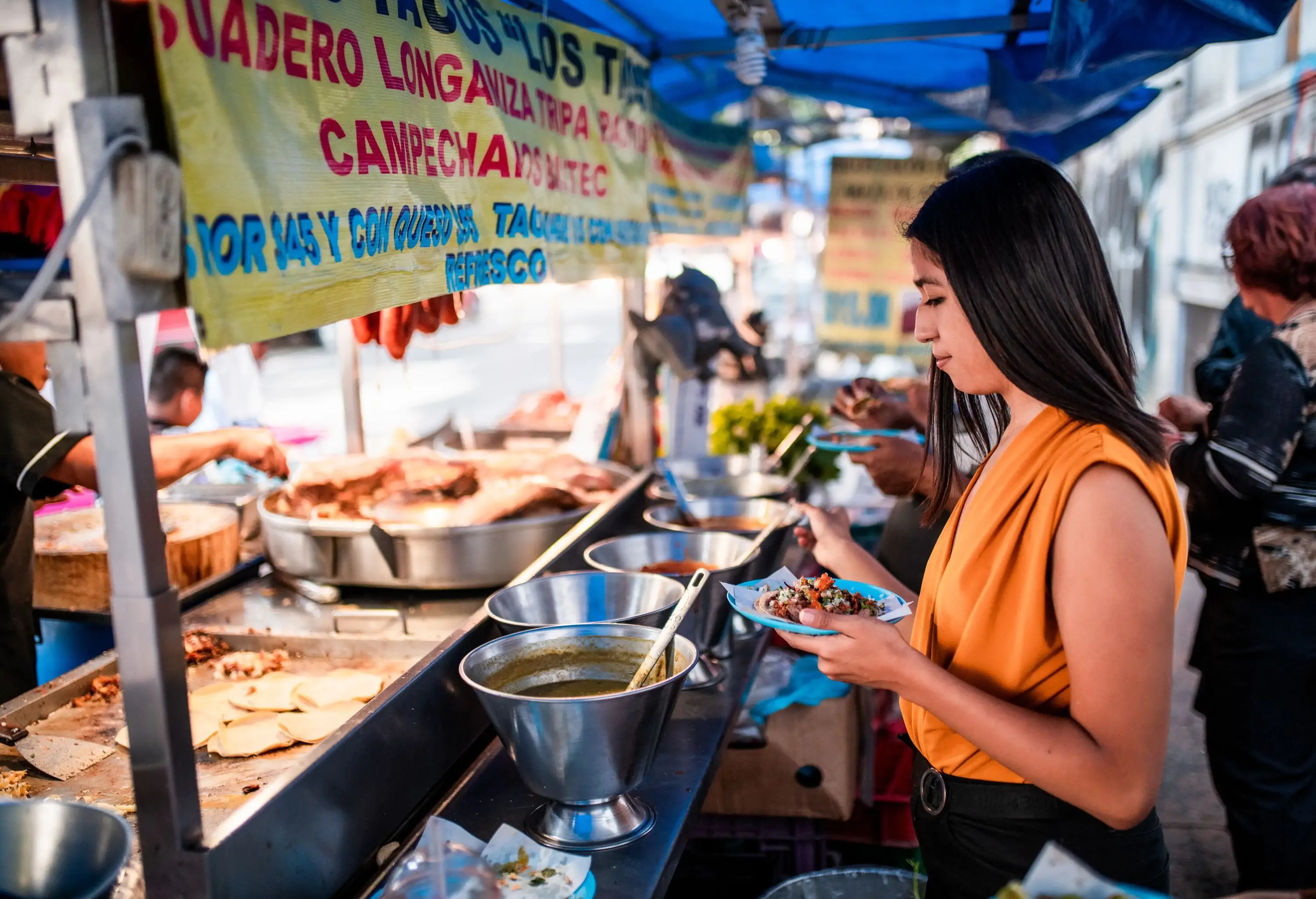 A woman, holding a plate of tacos, carefully selects a sauce from an enticing array displayed in front of a food stall.