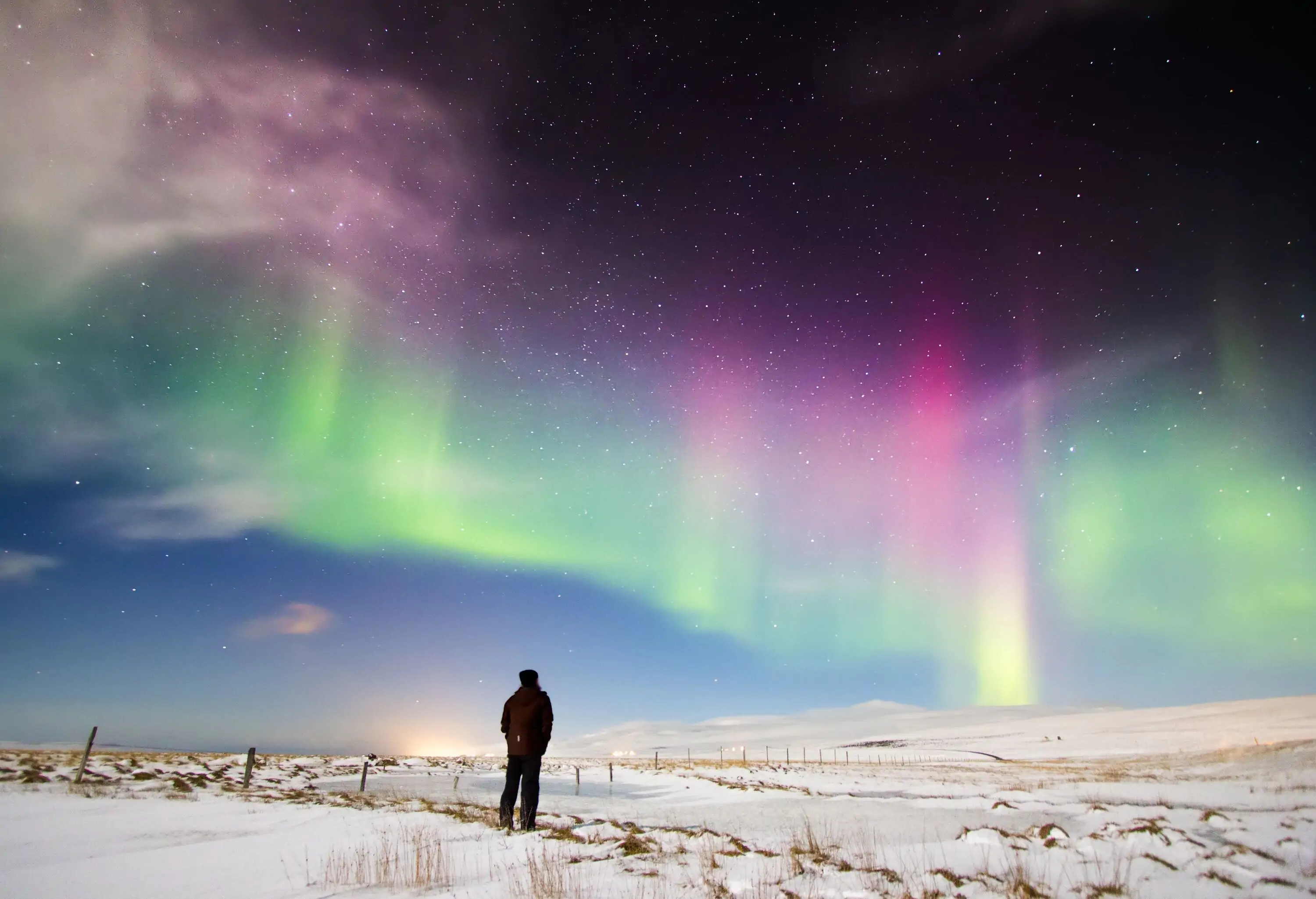 A man stands in awe, gazing at the vibrant colours of the Aurora Borealis dancing across a starry blue sky, with snow-covered ground beneath his feet.