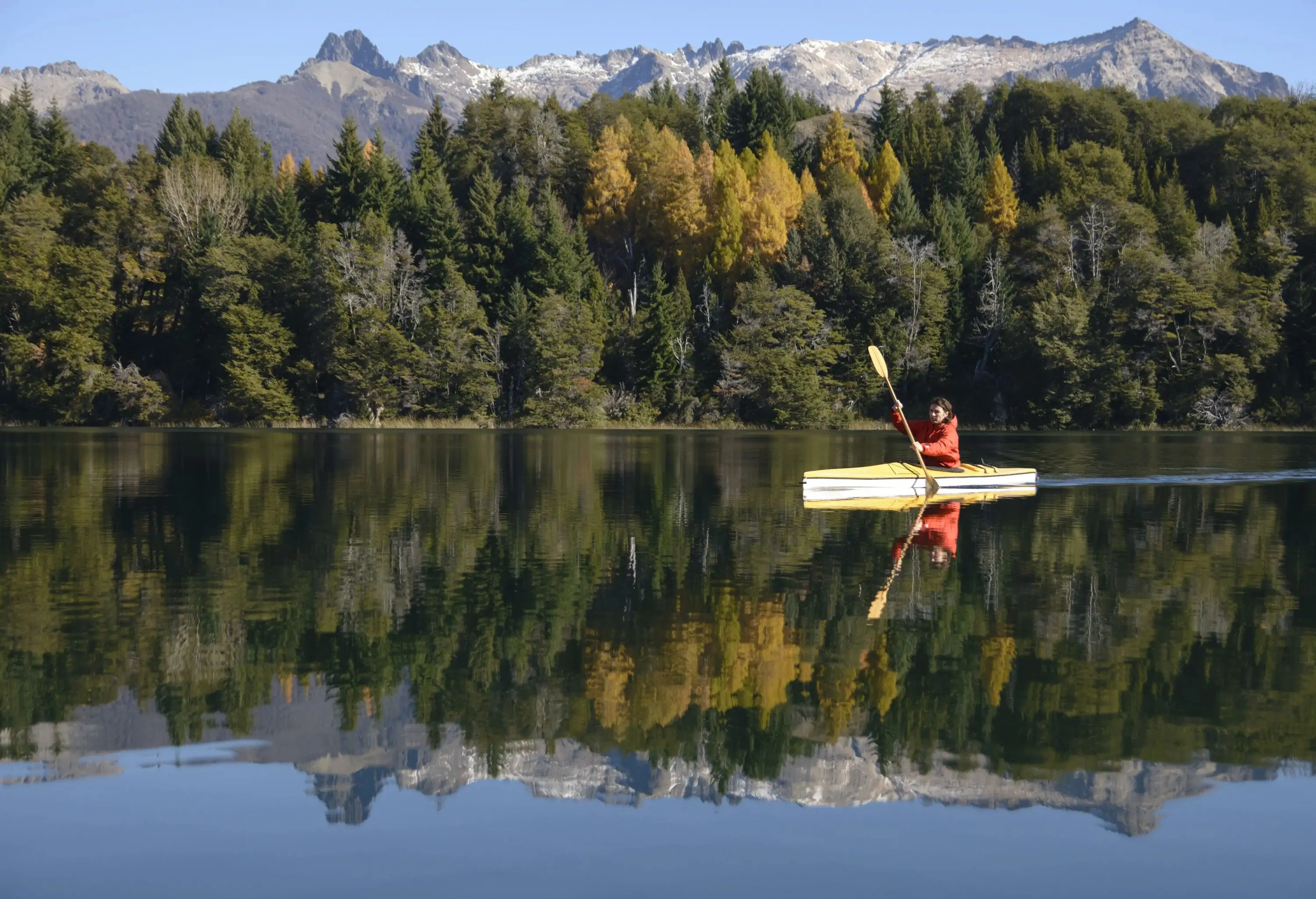 A person on a kayak paddling across a river surrounded by dense trees and mountains.