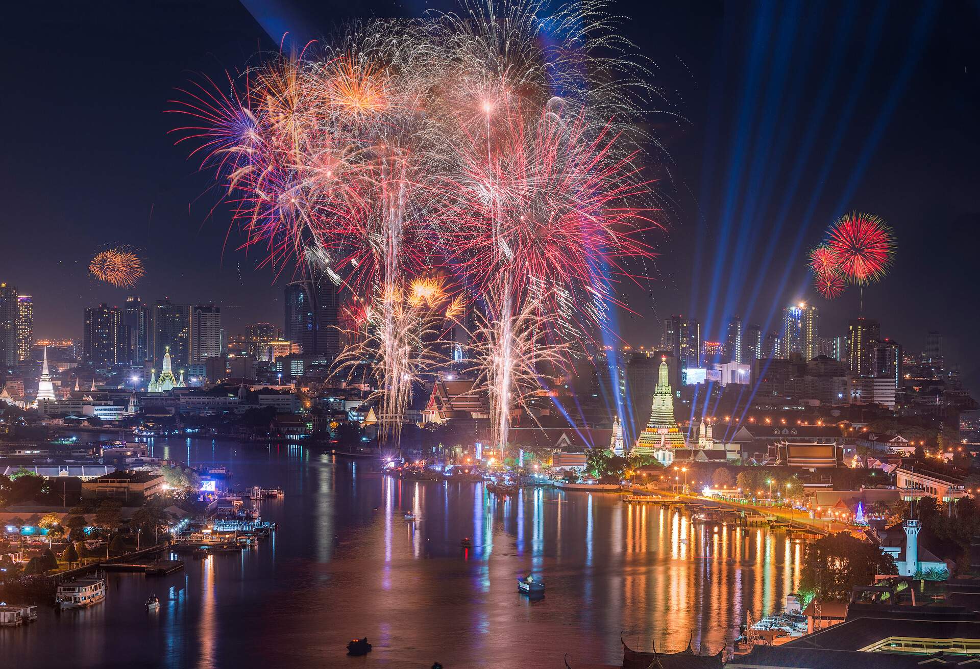 Colourful fireworks explode in the night sky above the brightly lit riverside cityscape and high-rise buildings.