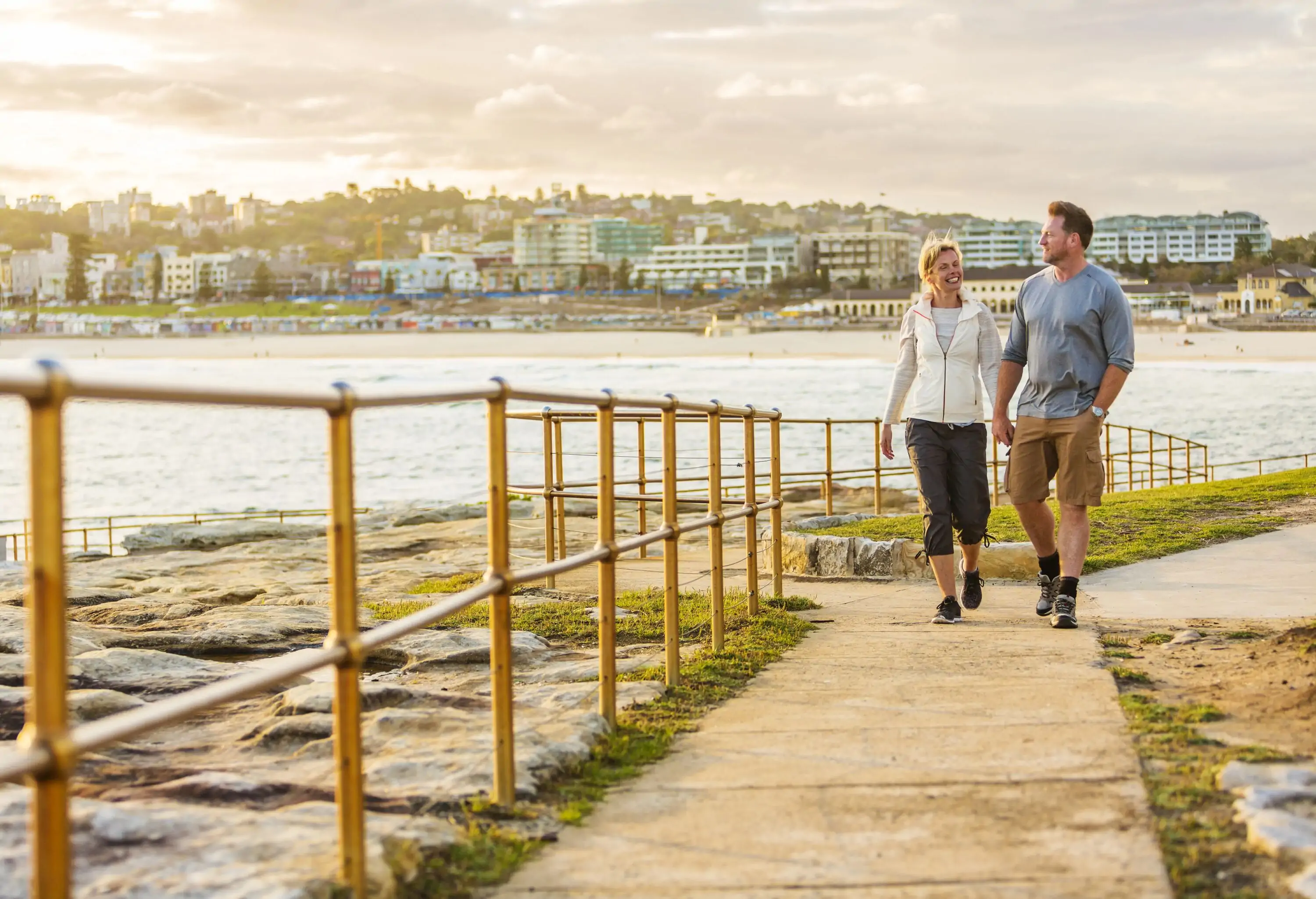 A couple enjoying the sunset while walking hand in hand on a seaside paved footpath with a view of an urban scenery in the background.