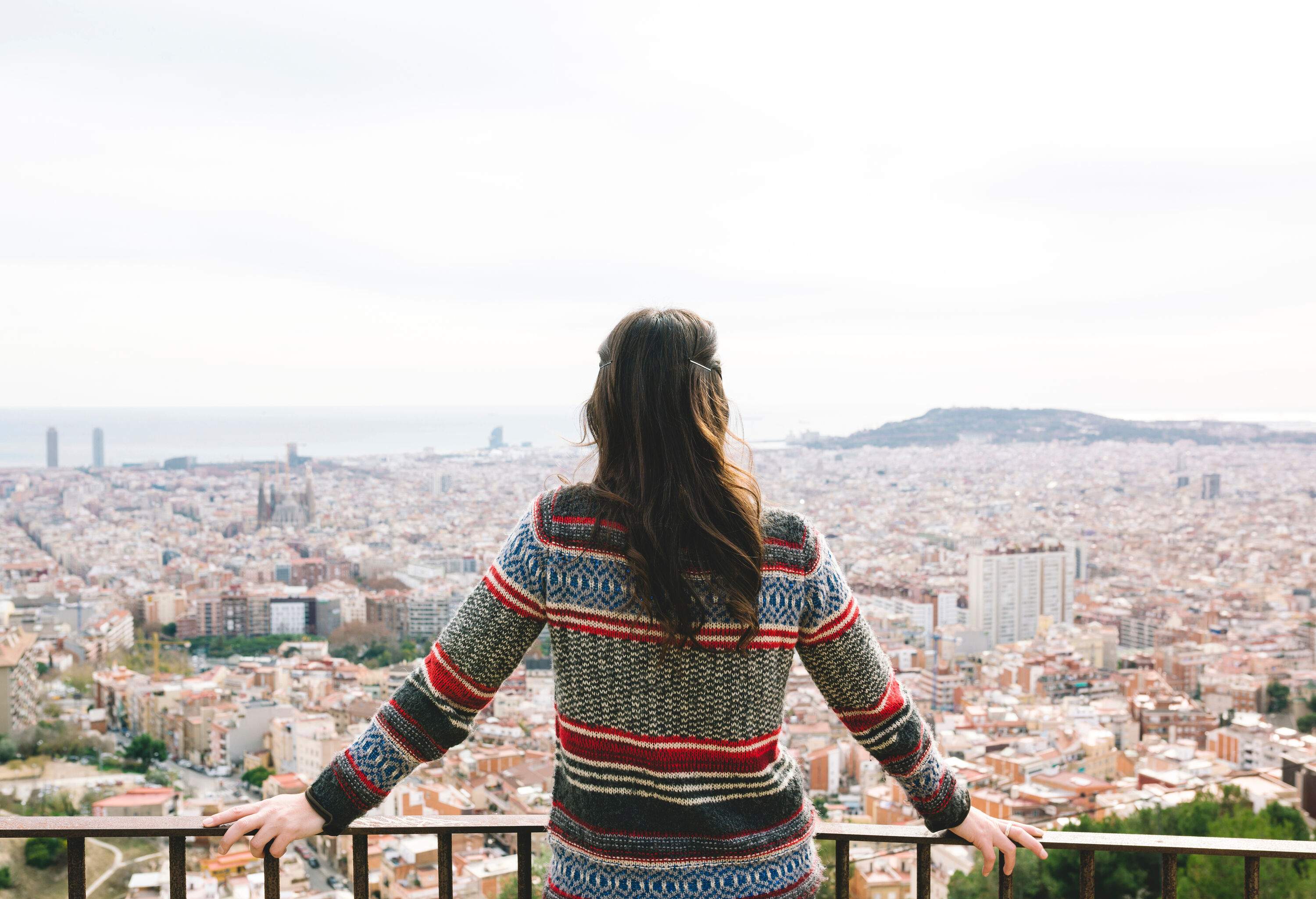 A woman in a sweater leaning on the railing of a balcony and taking in the cityscape.