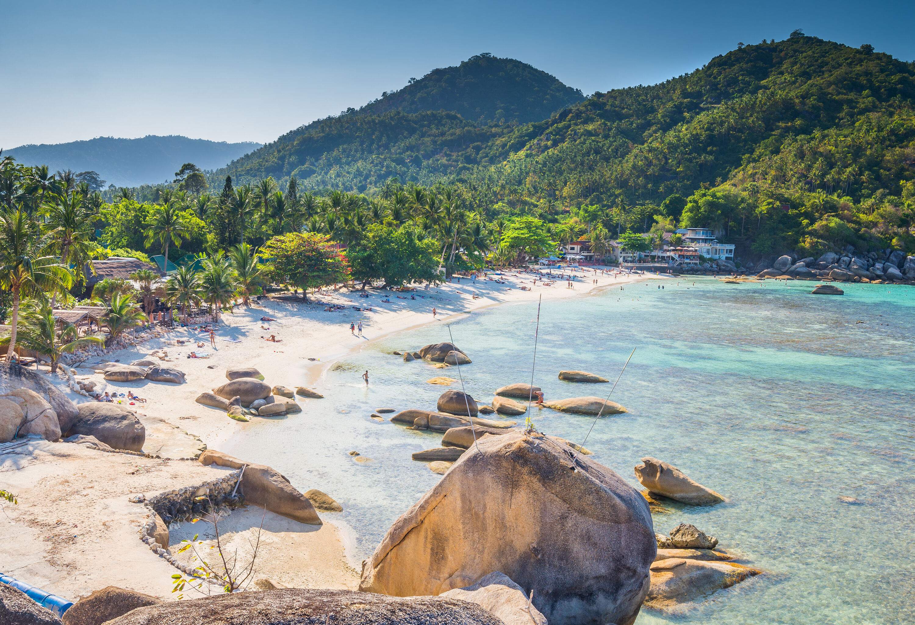 A picturesque view of a tranquil beach with boulders and a white sand shore lined with resort buildings surrounded by tall green trees beneath the forested mountain range.