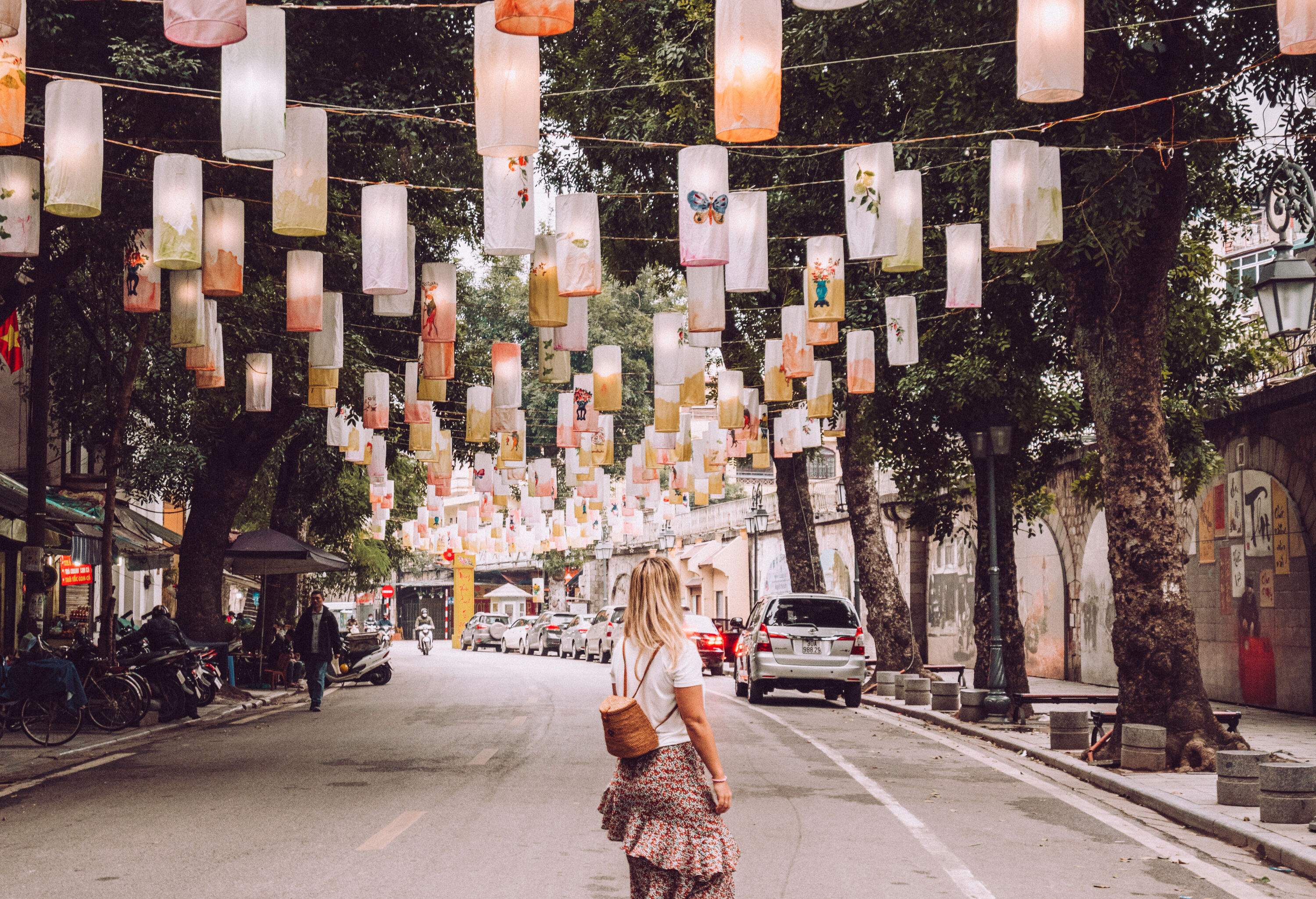 A woman standing in the middle of a street with cylindrical lanterns hanging over it.