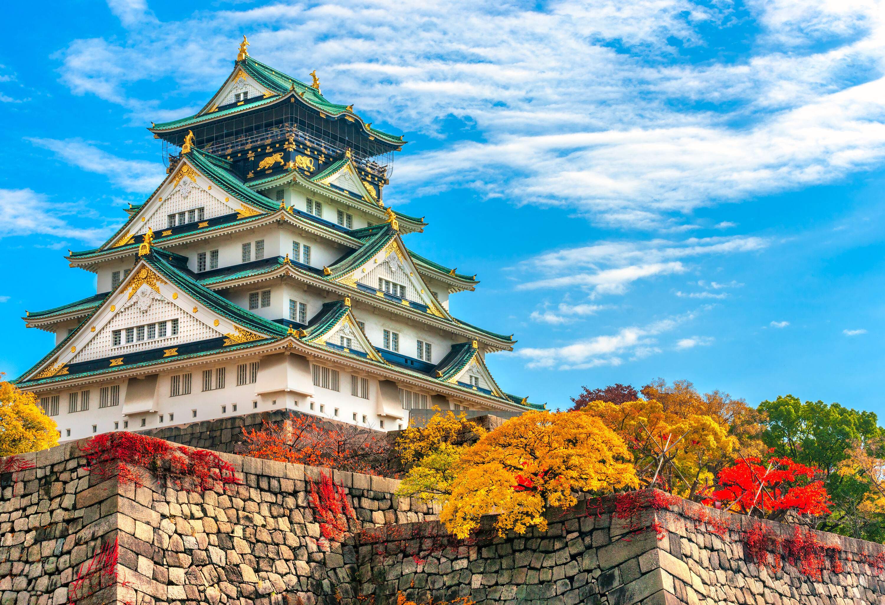 A Japanese castle encased in rough stone walls with colourful trees around it.