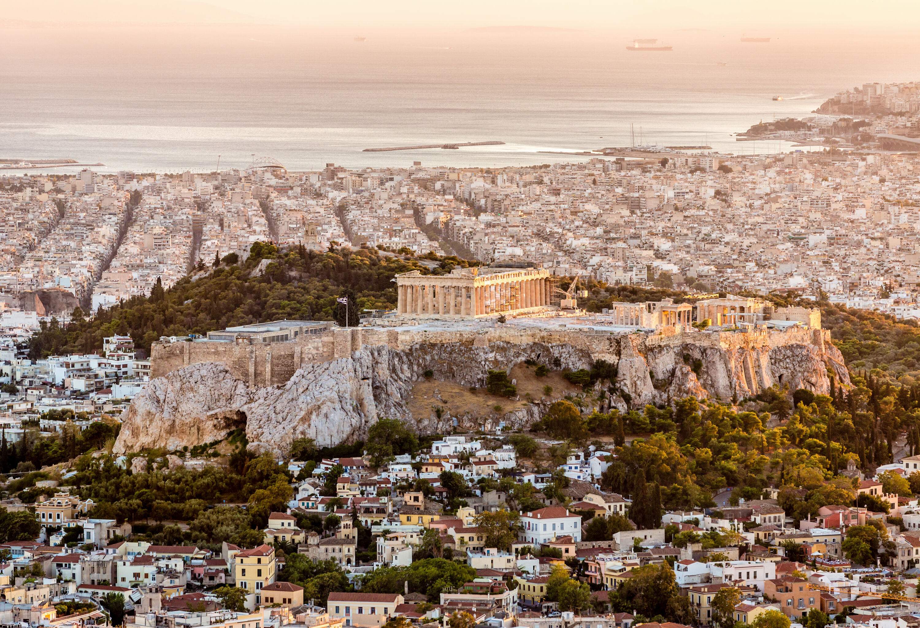 Aerial view of the city of Athens with the Acropolis in the middle of a cliff surrounded by tall green trees at sunset.