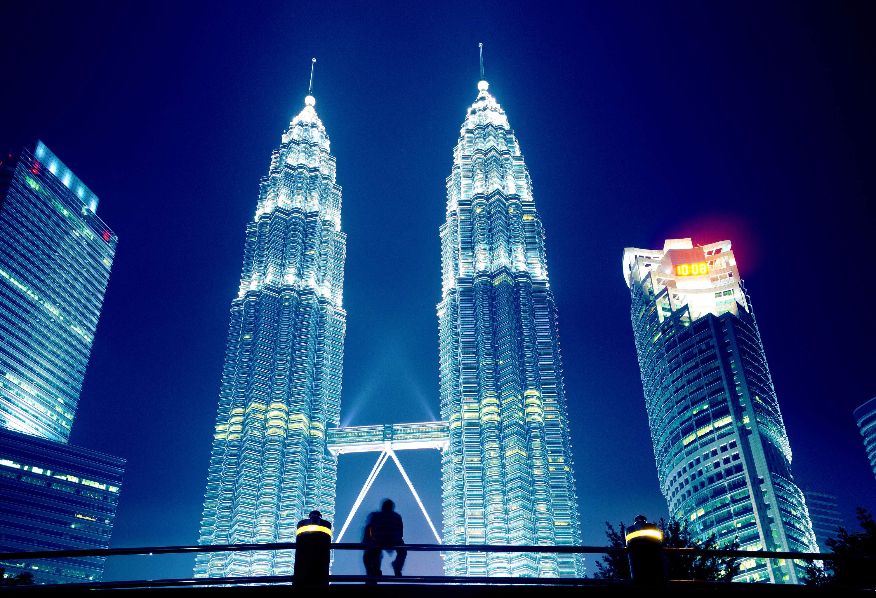 A man's silhouette sitting on a railing, gazing at the luminous Petronas Twin Towers against the night sky.