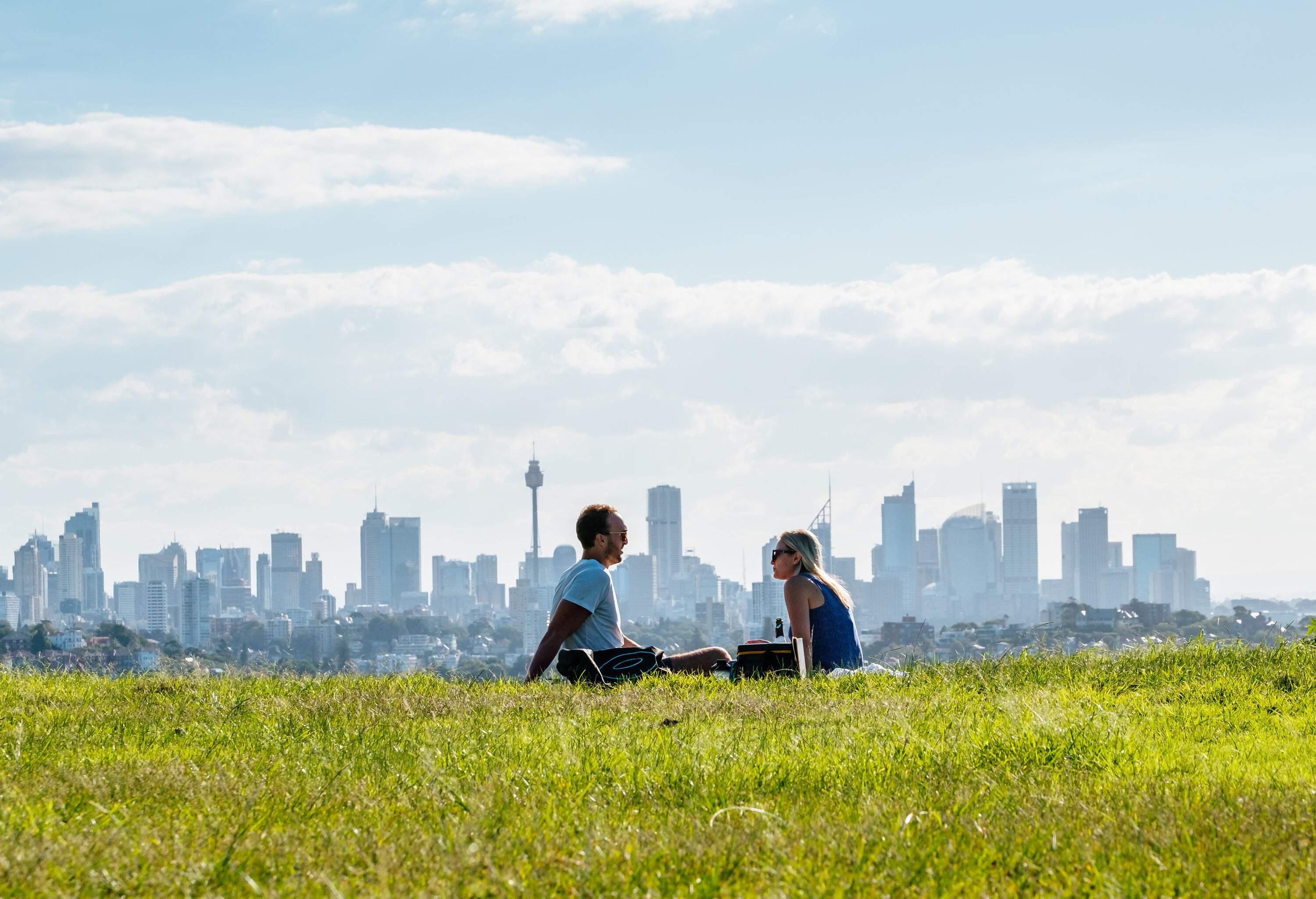 A couple enjoying the sunlight while sitting on a lush landscape overlooking the modern urban cityscape against the cloudy sky.