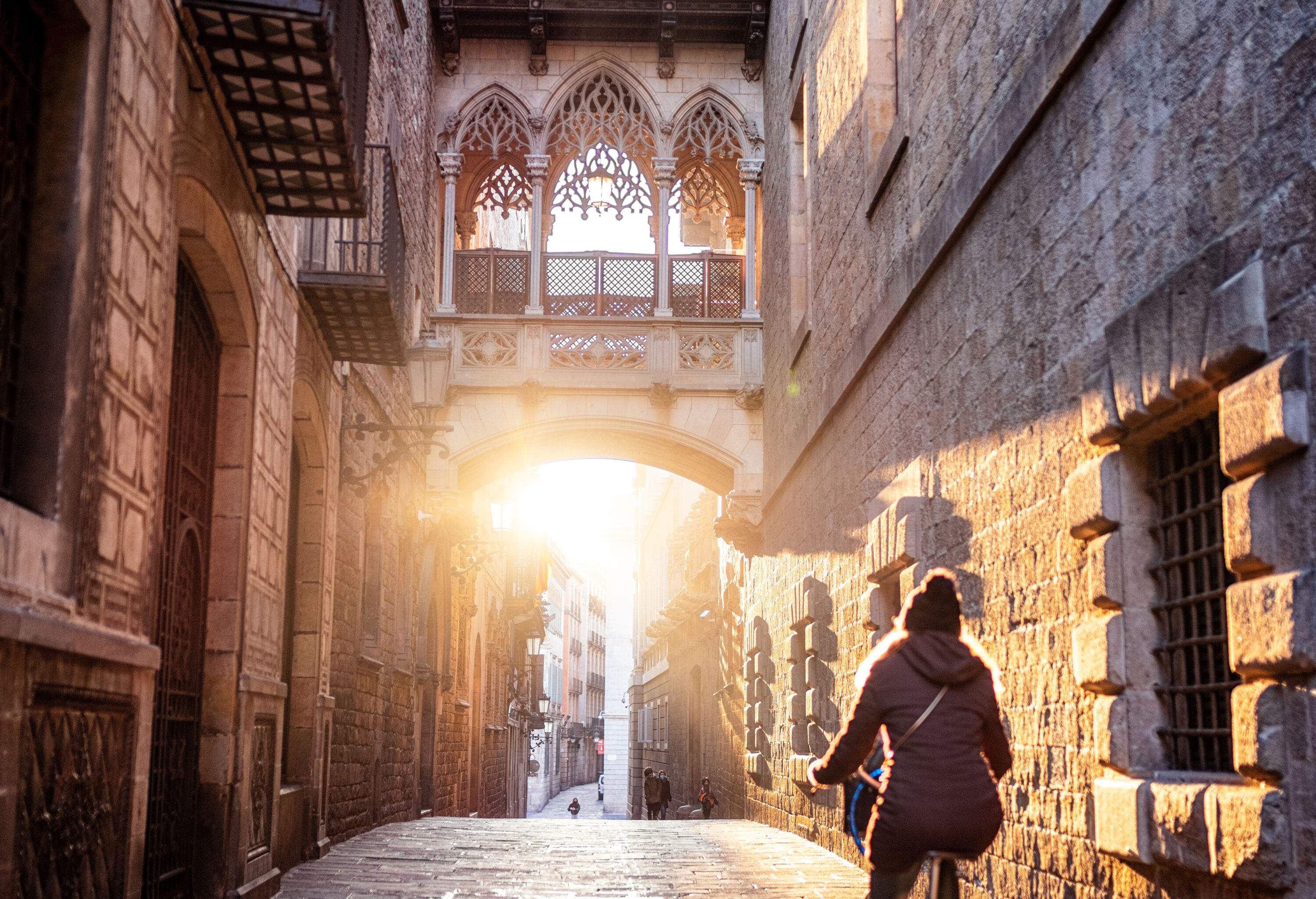 A woman pedals along a cobbled street lined with Gothic buildings at sunrise.