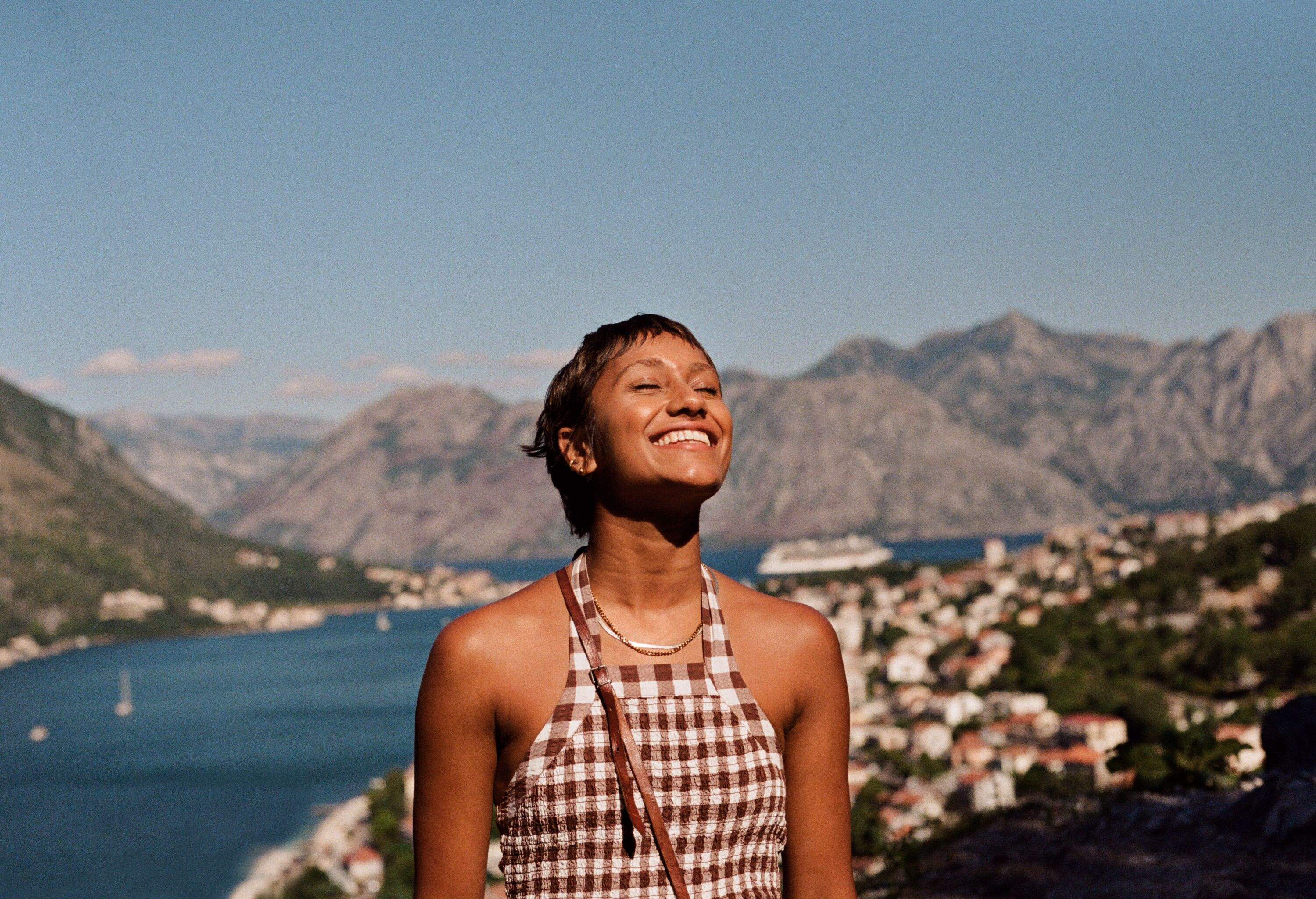 Smiling young woman enjoying sunlight against mountain range and sea during vacation