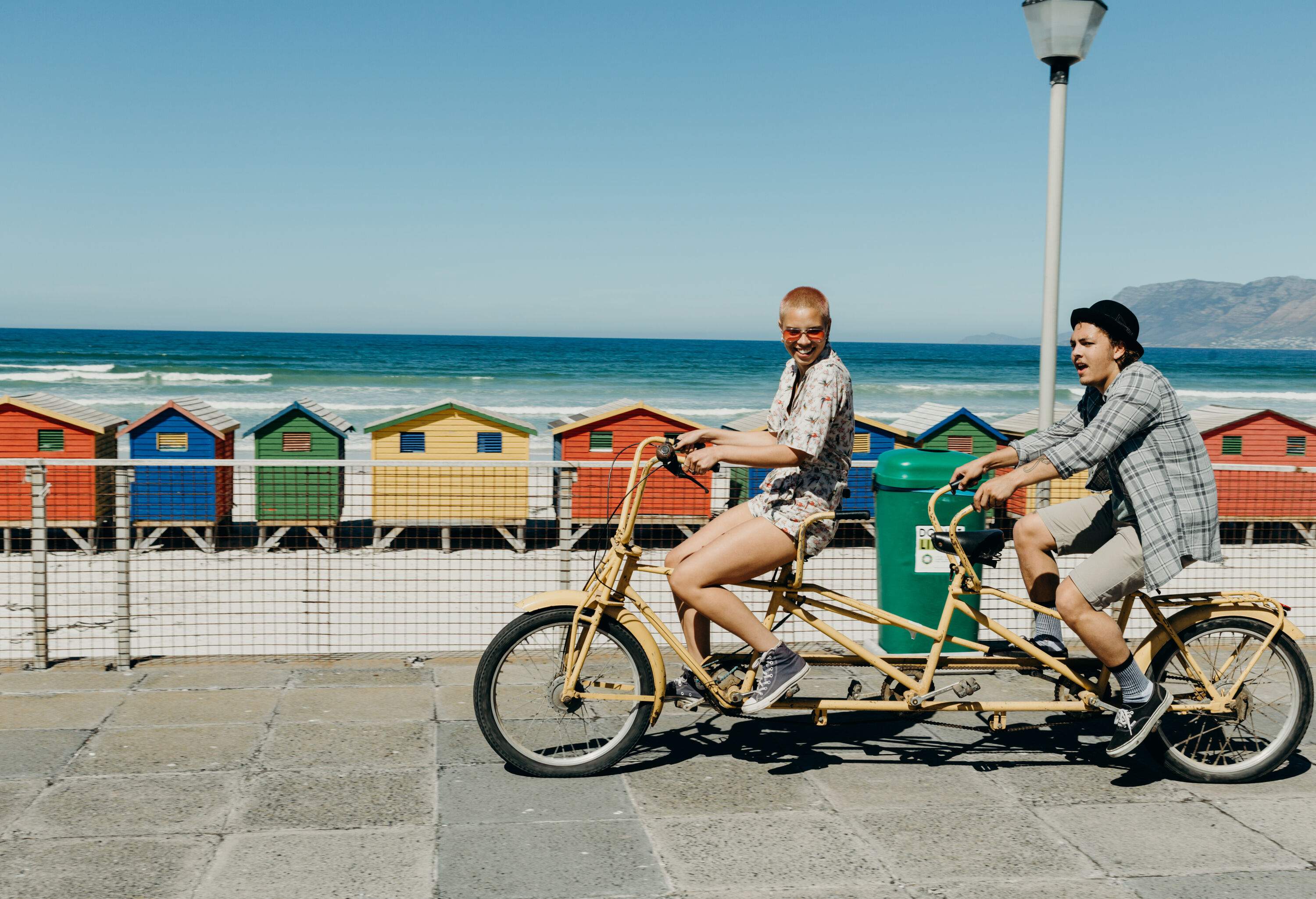 Two friends in casual clothes ride a tandem bicycle on a promenade beside a beach with a row of colourful beach huts.