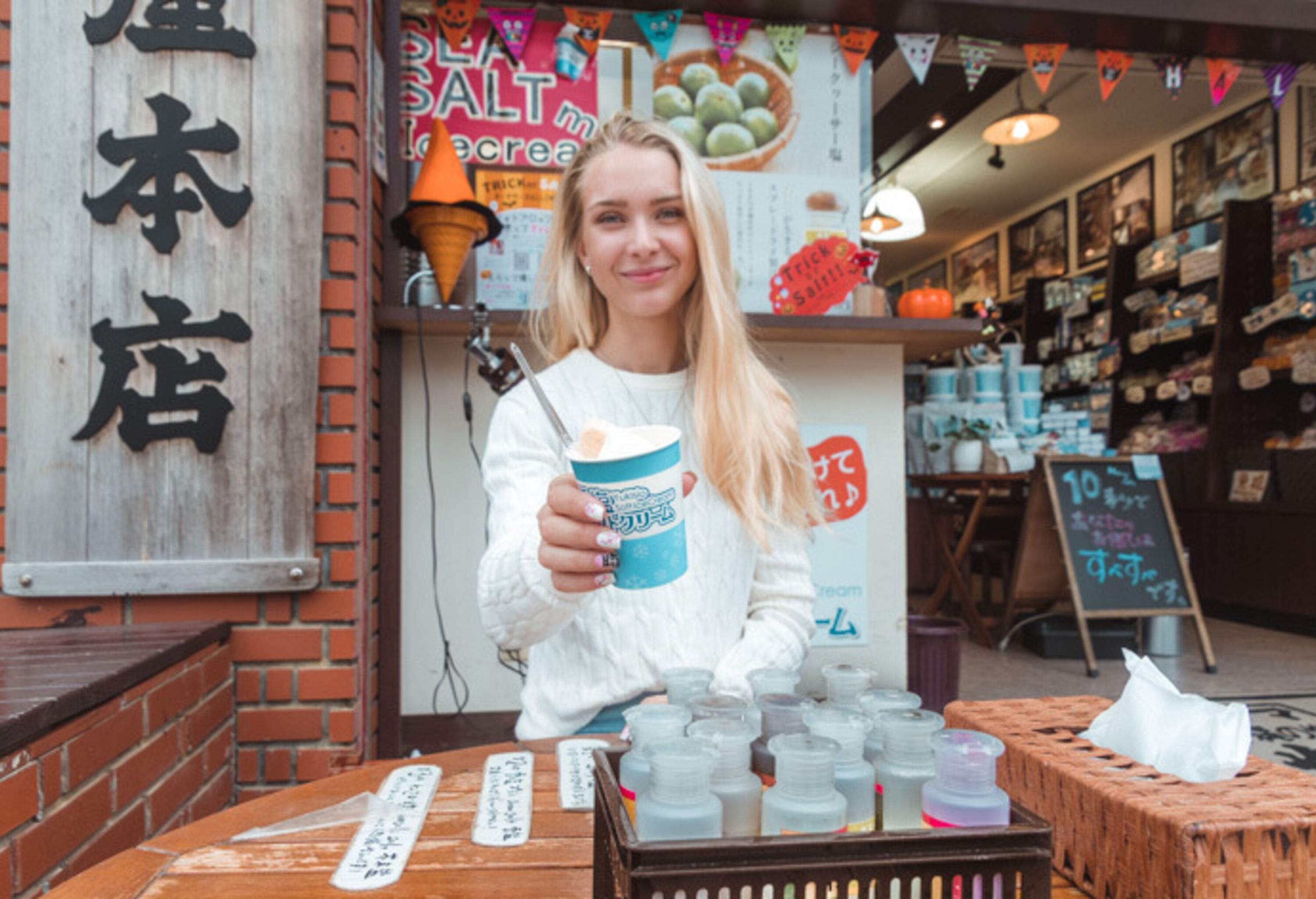 A long-haired blonde pretty lady shows off a cup of ice cream while sitting outdoors on a wooden table.