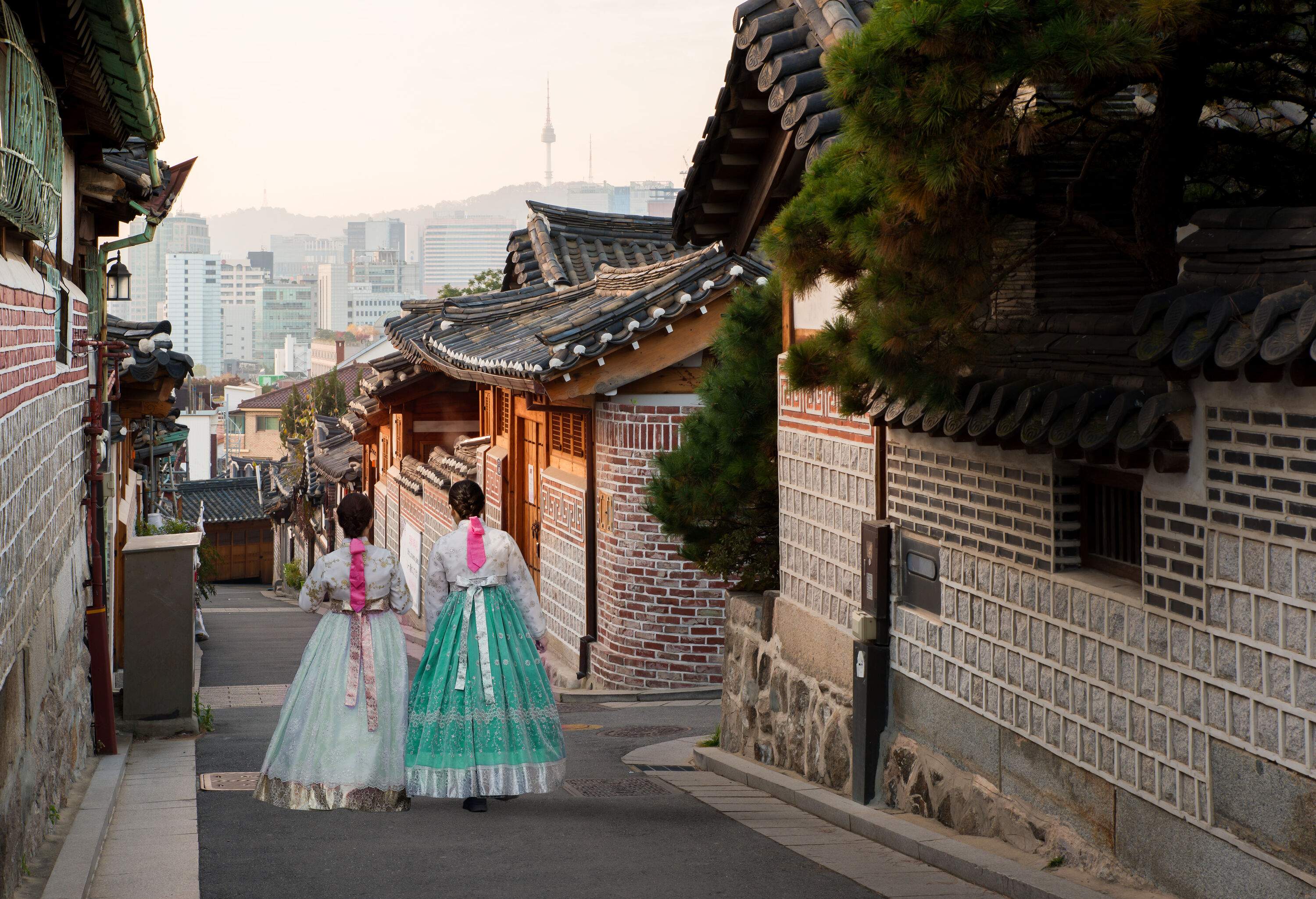 Back view of two women elegantly dressed in hanbok, strolling through a charming neighbourhood of traditional-style houses.