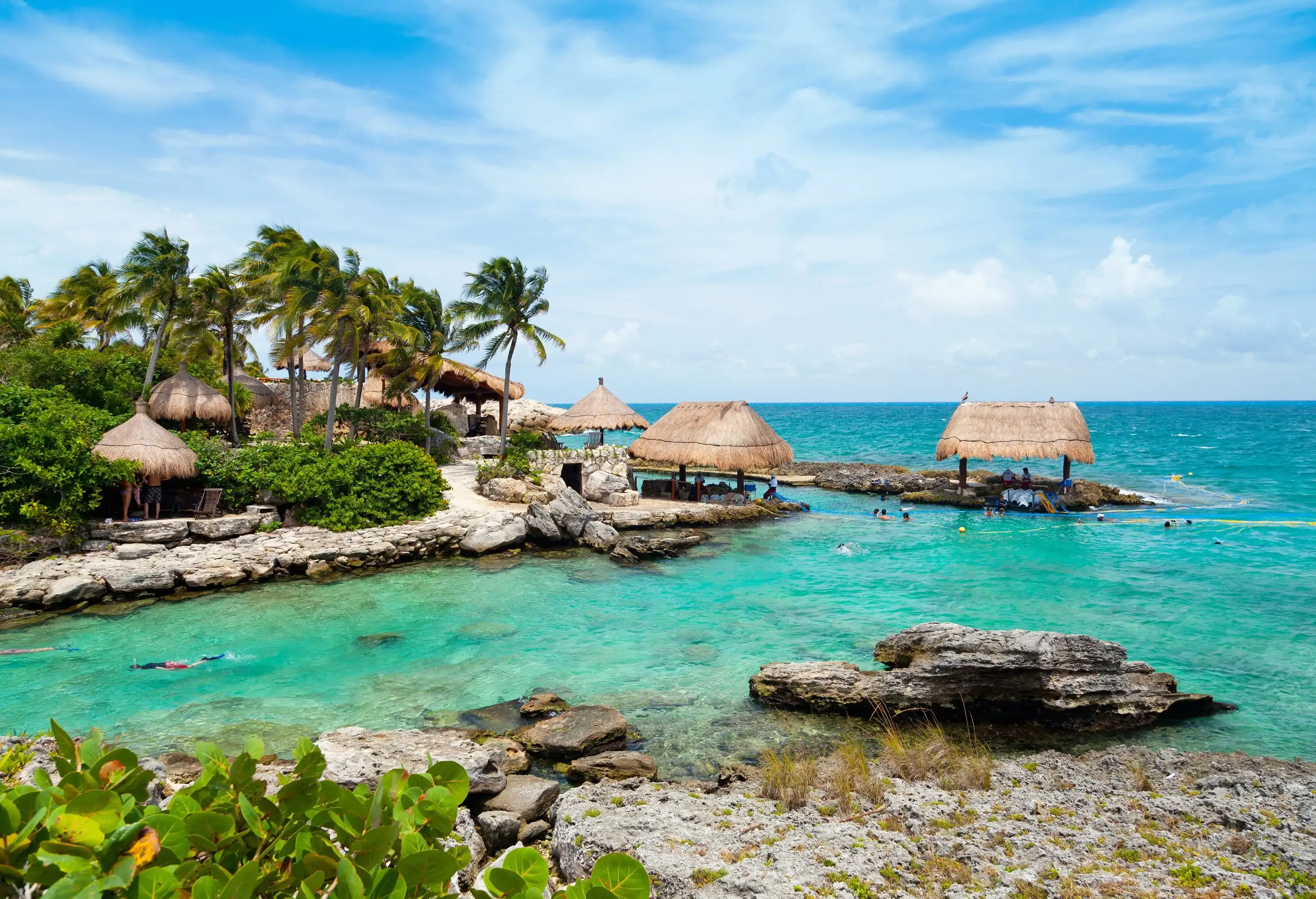 Wooden beach huts on the rocky shore surrounded by tall green trees by the turquoise beach.