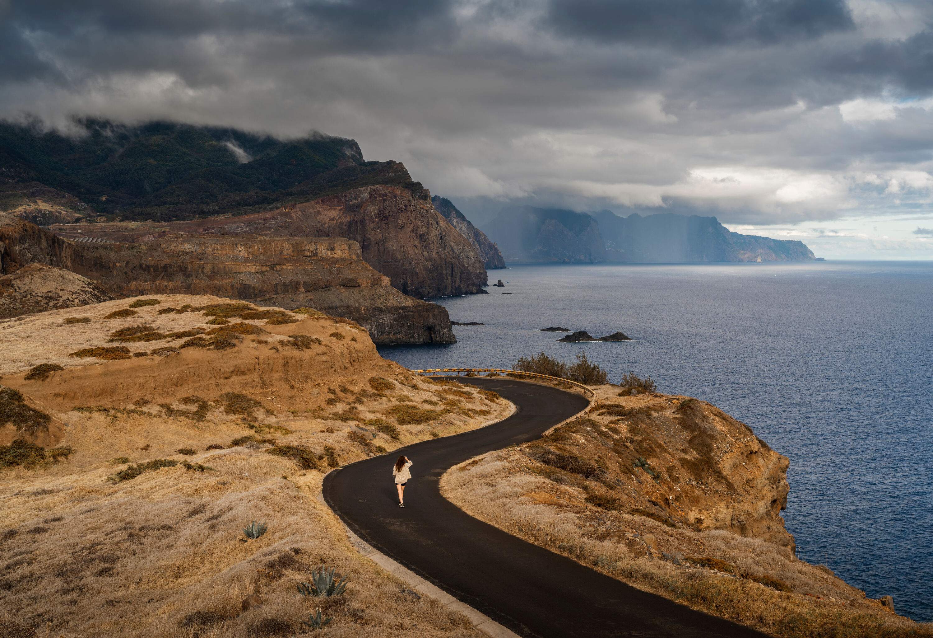 A lone woman walking on a winding road across a barren seaside cliff.