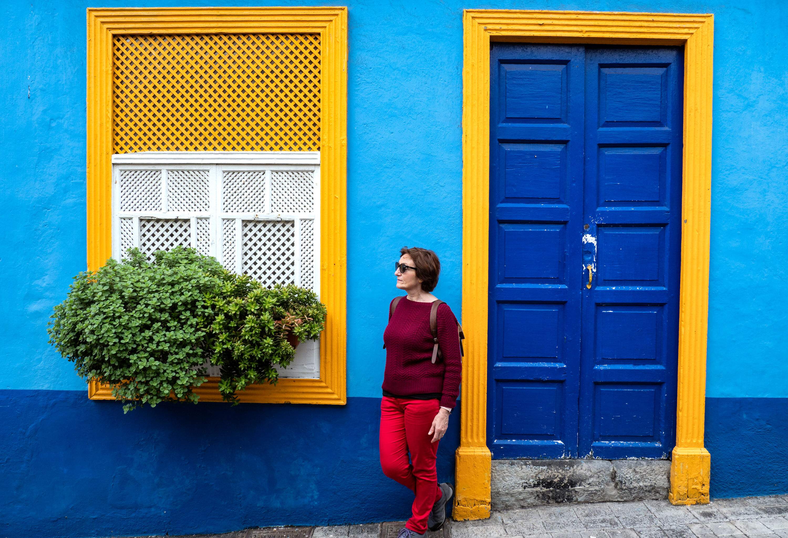 A casually dressed person stands between the window and door of a house painted blue.