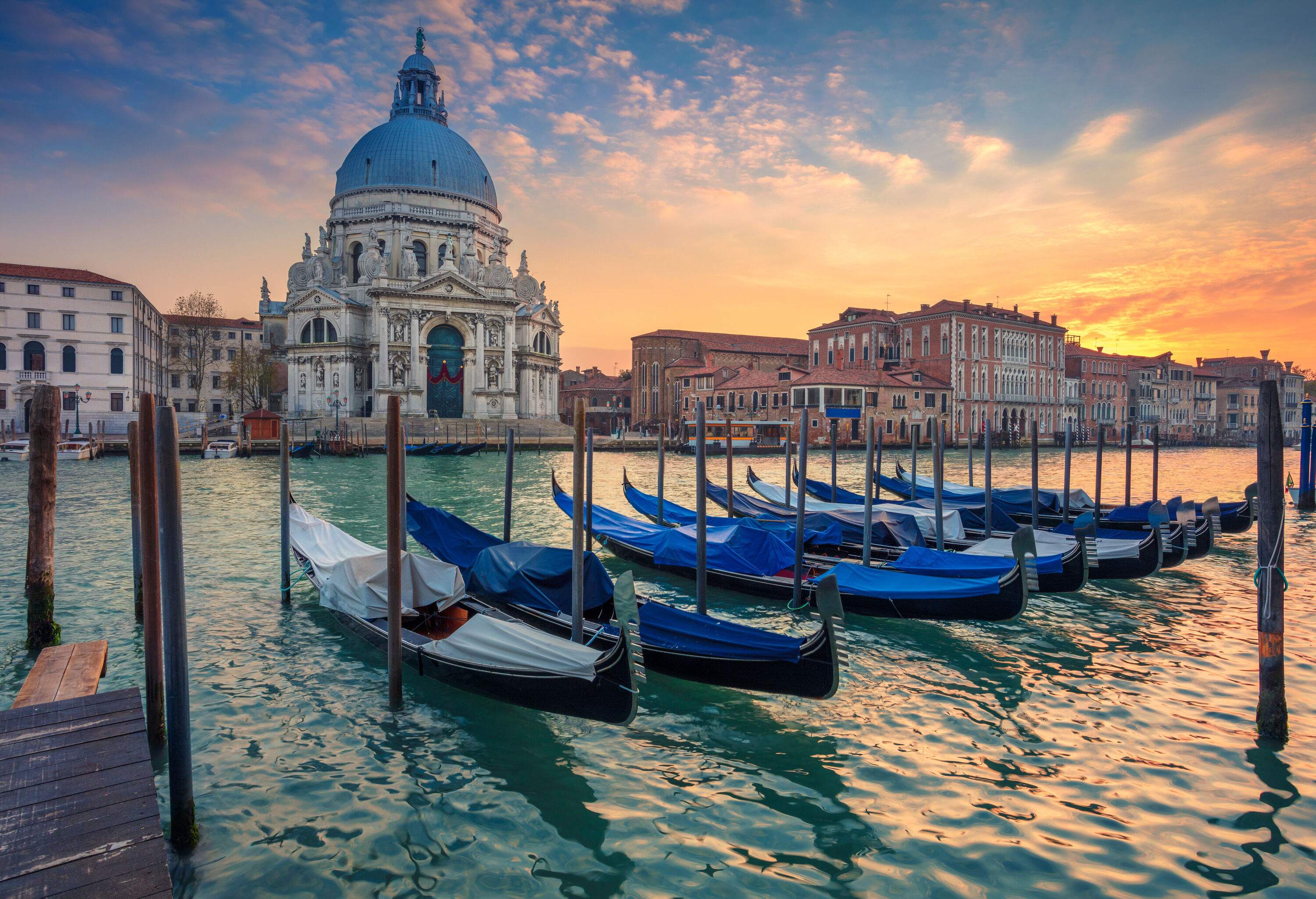 A row of gondolas anchored on wood pilings in a harbour with views of Santa Maria della Salute, a domed Baroque church beneath an orange sky.