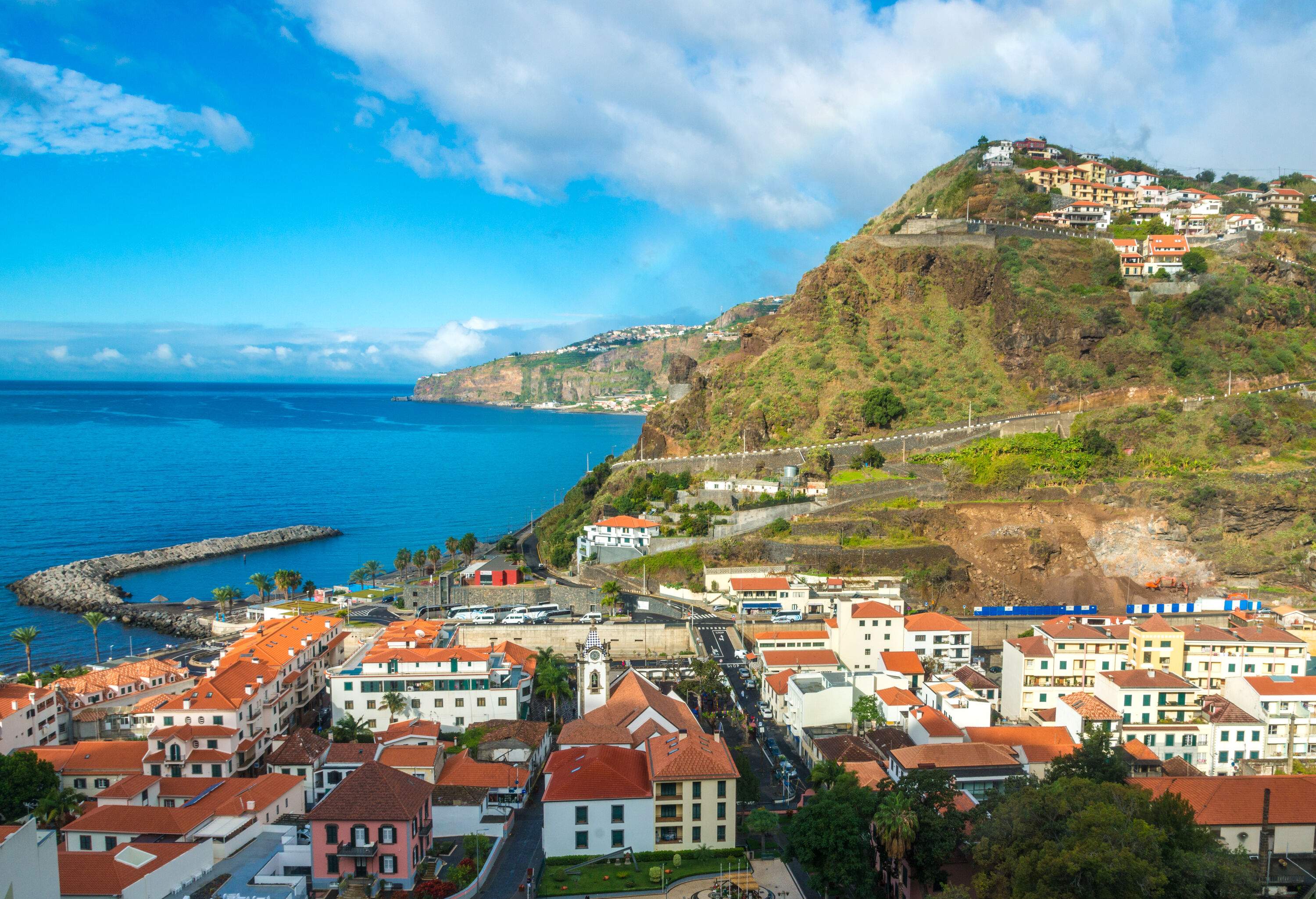 Skyline view of a coastal town of red brick-roofed buildings on the valley and hilltop overlooking the blue sea against the cloudy blue sky.