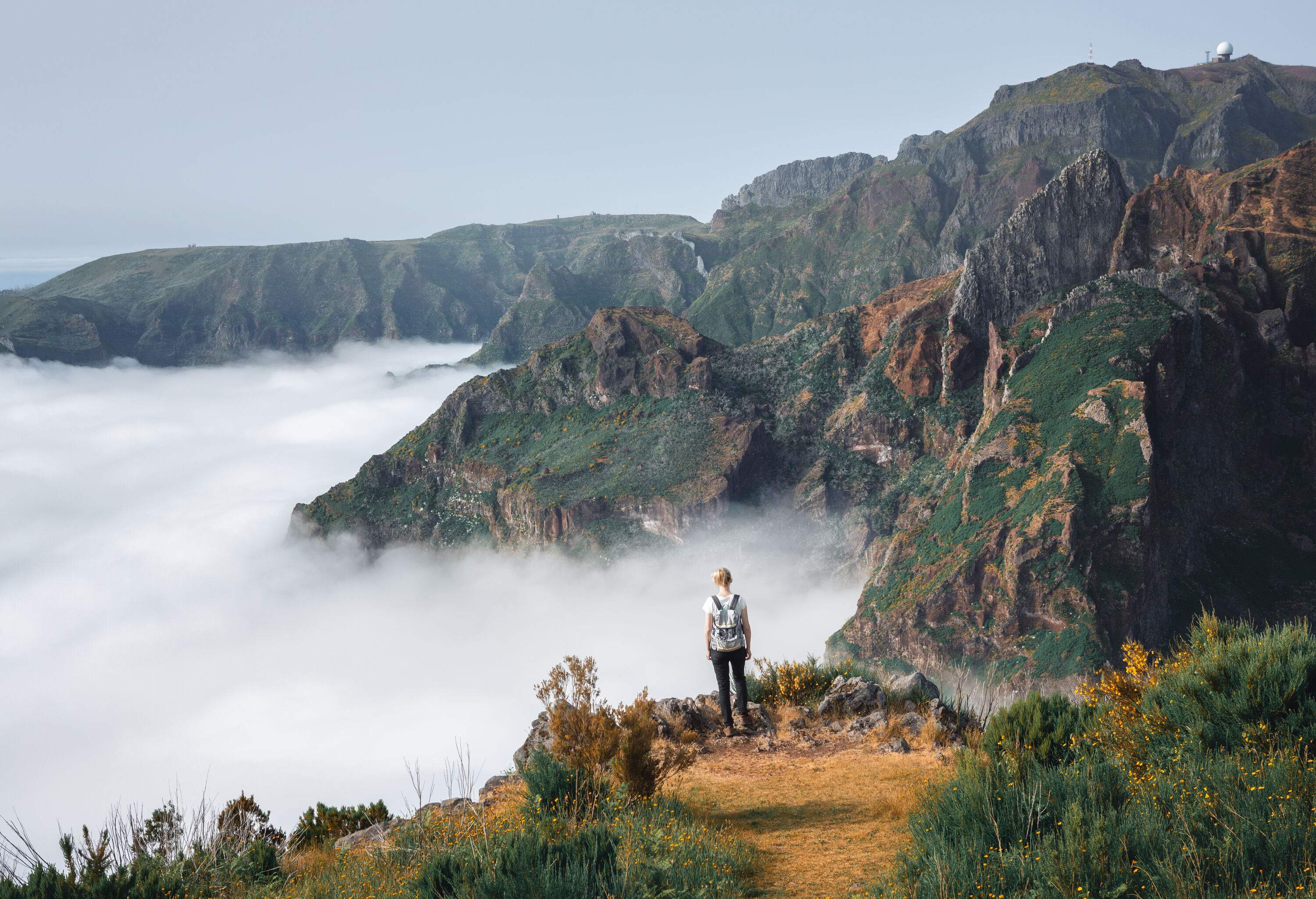 A hiker standing on the brink of a cliff, staring down over a sea of clouds below the mountains.