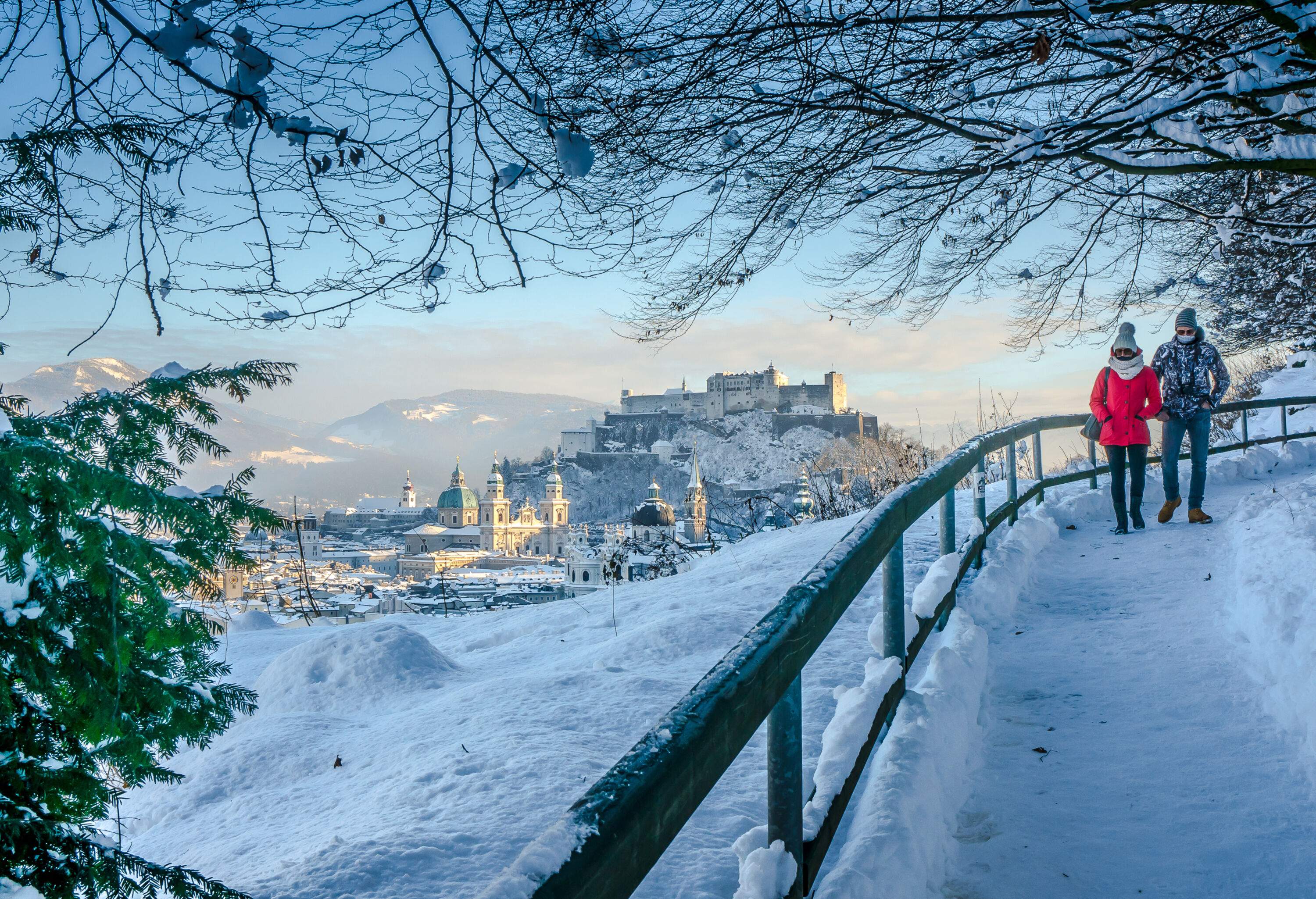 A couple of people walking down a snow-covered walkway with a view of old town structures in the background.