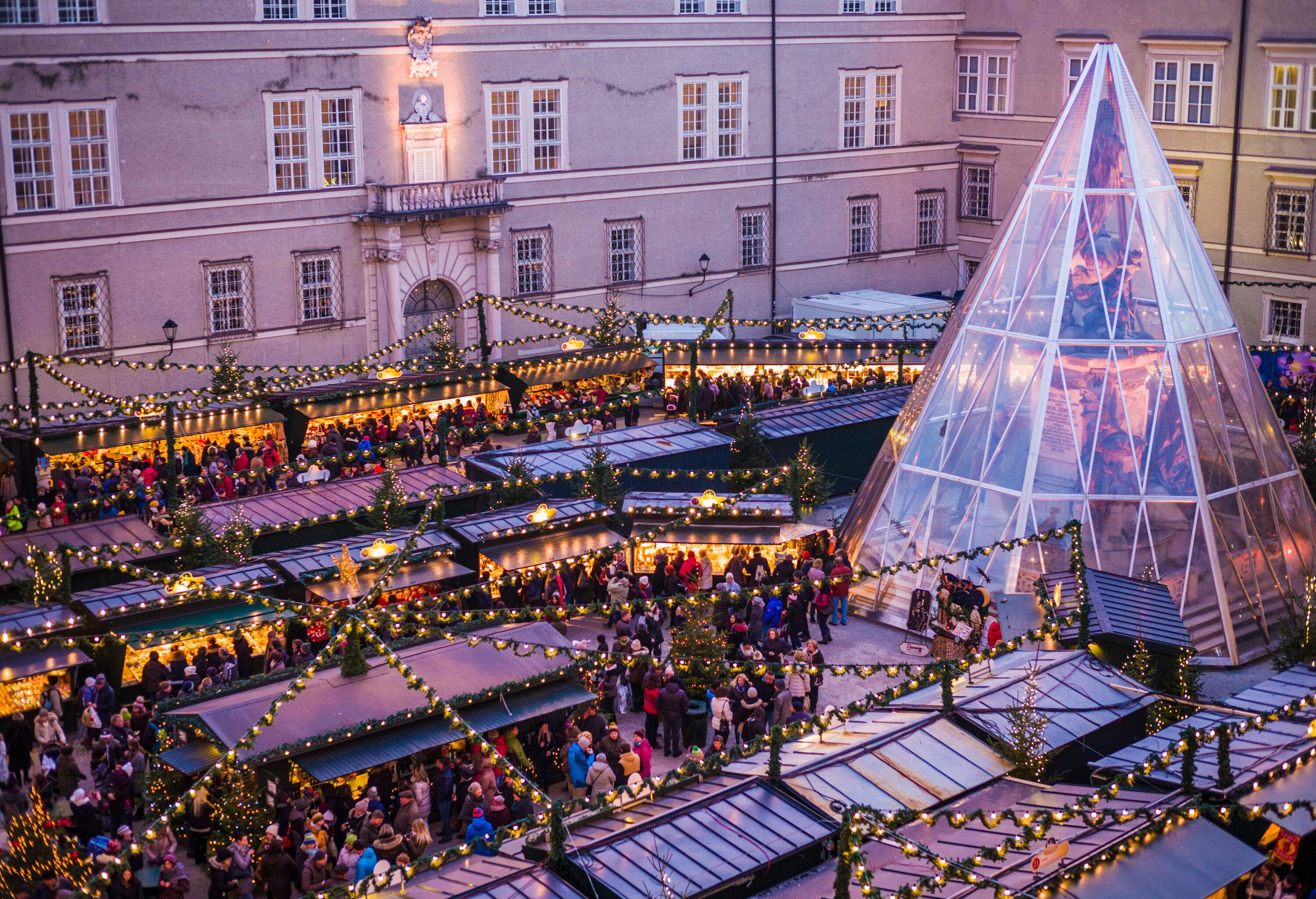 A crowded cathedral square with a lively Christmas market and a glass pyramid housing a statue.