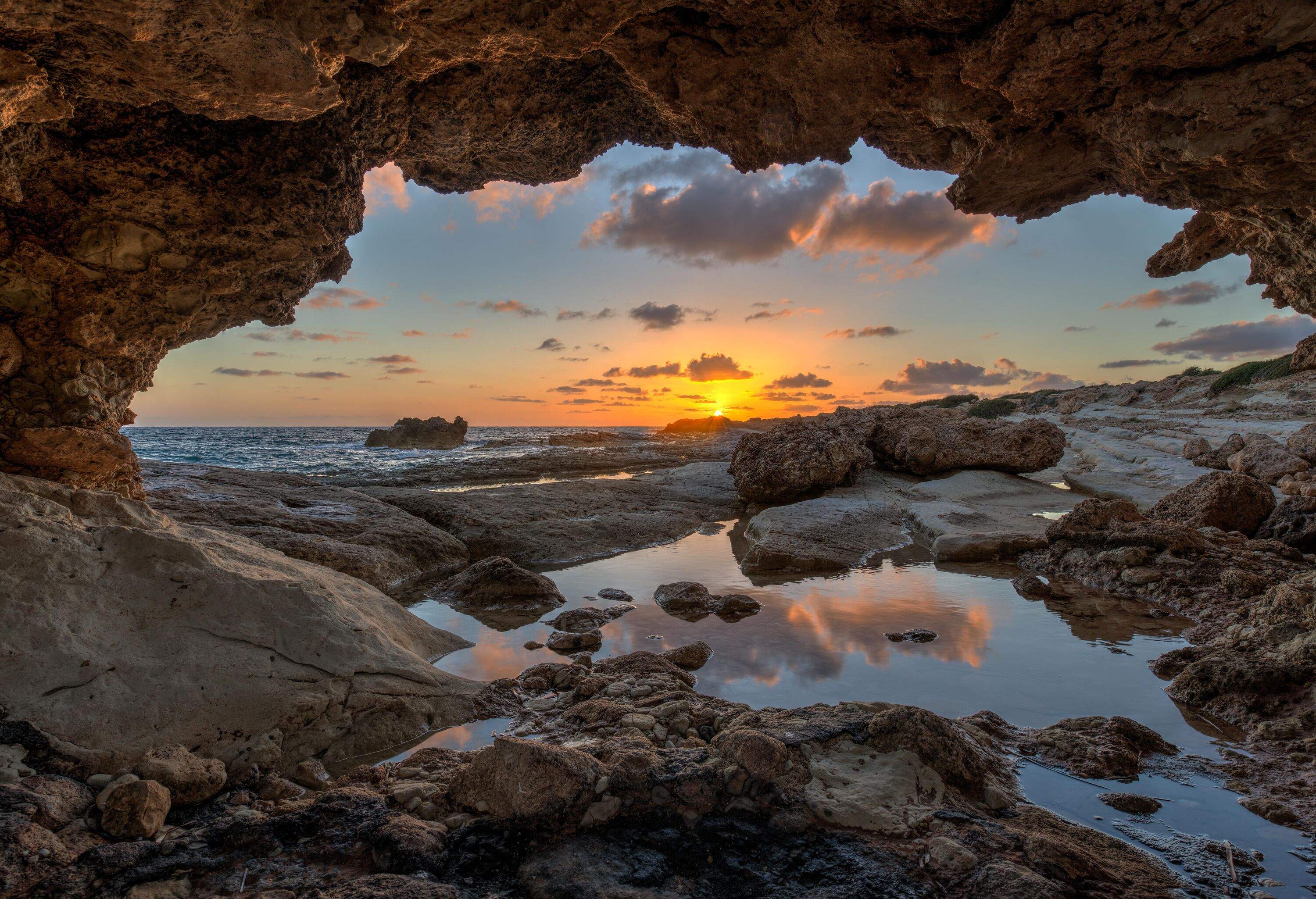 A picturesque view of the rocky shore of a serene sea, as seen from the opening of a cave, captured during the enchanting sunset.