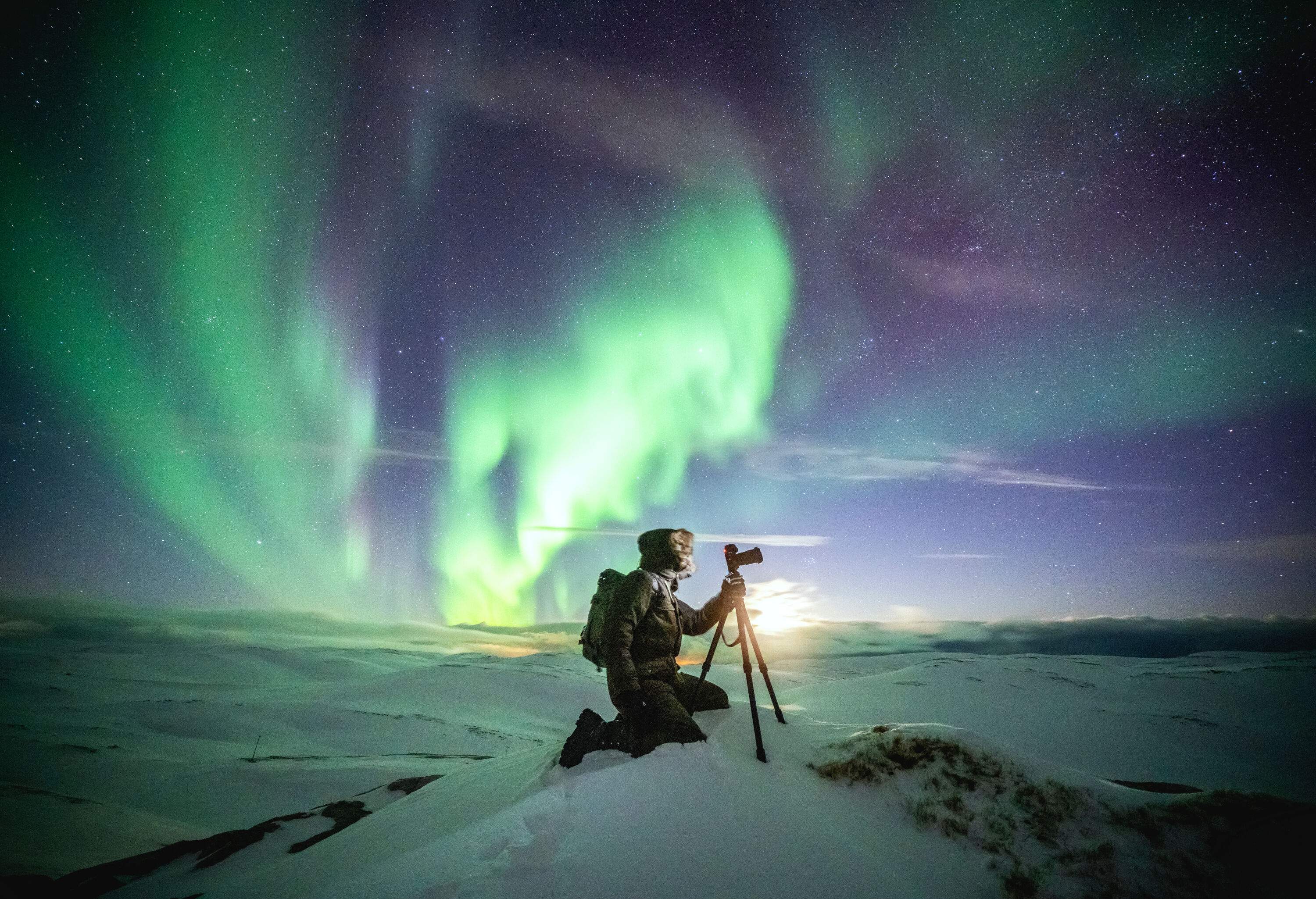 A photographer takes pictures of the Northern green lights dazzling in the sky with bended knees on his tripod.