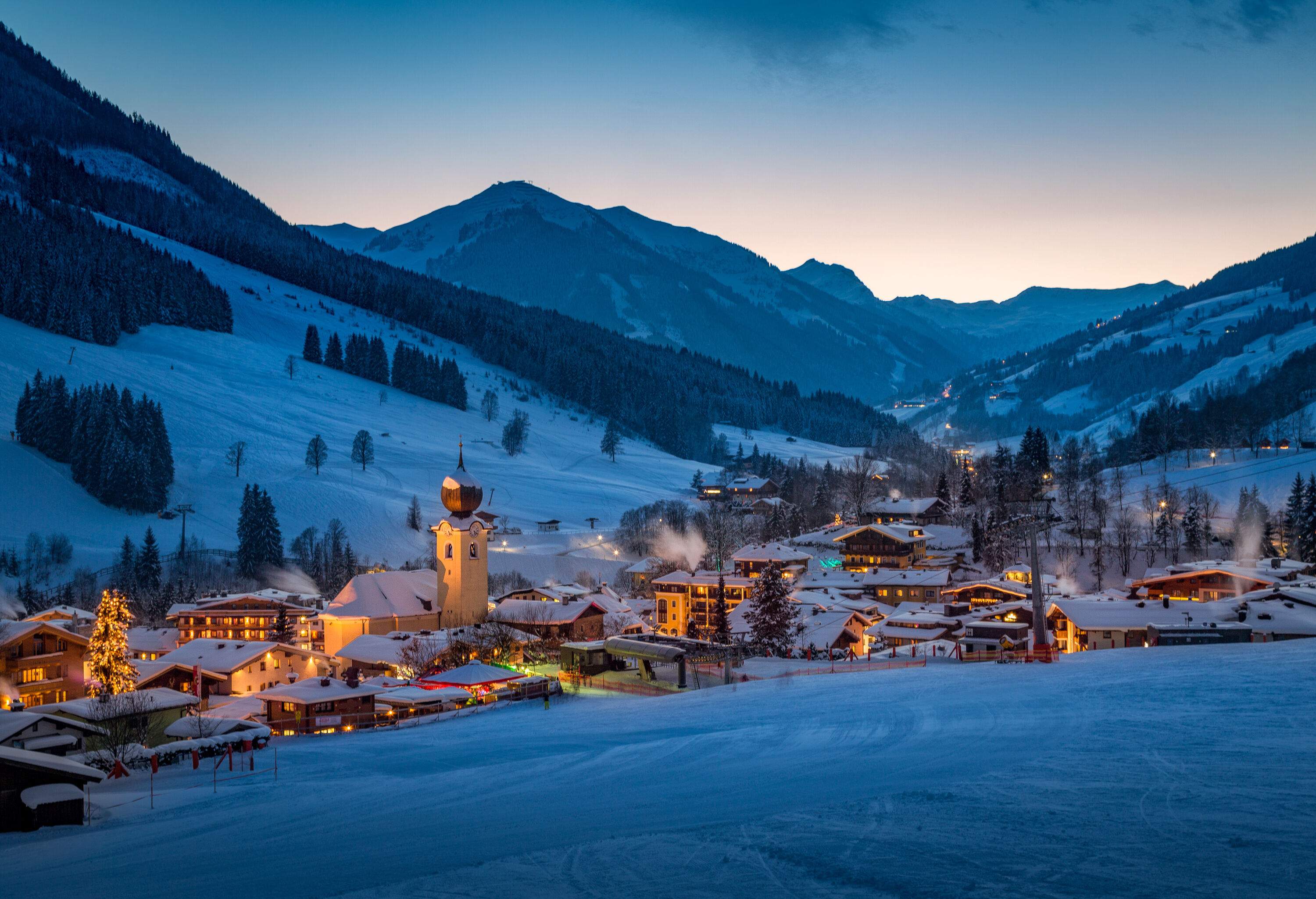 Illuminated village at night in a rolling forested mountain valley covered in deep snow.