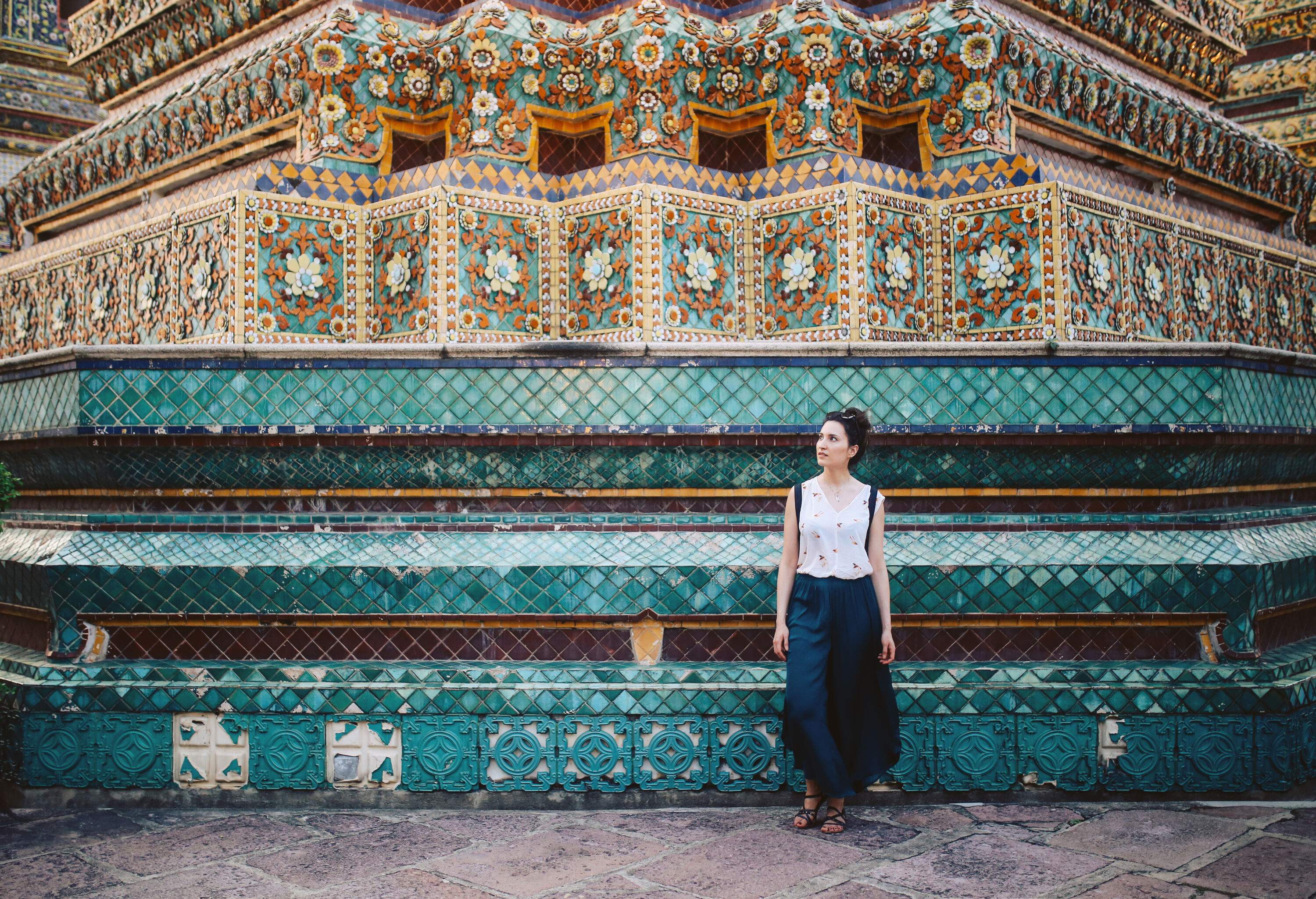 A woman wearing a navy blue sarong and white shirt stands next to a terraced structure decorated with ceramic tiles with floral patterns.