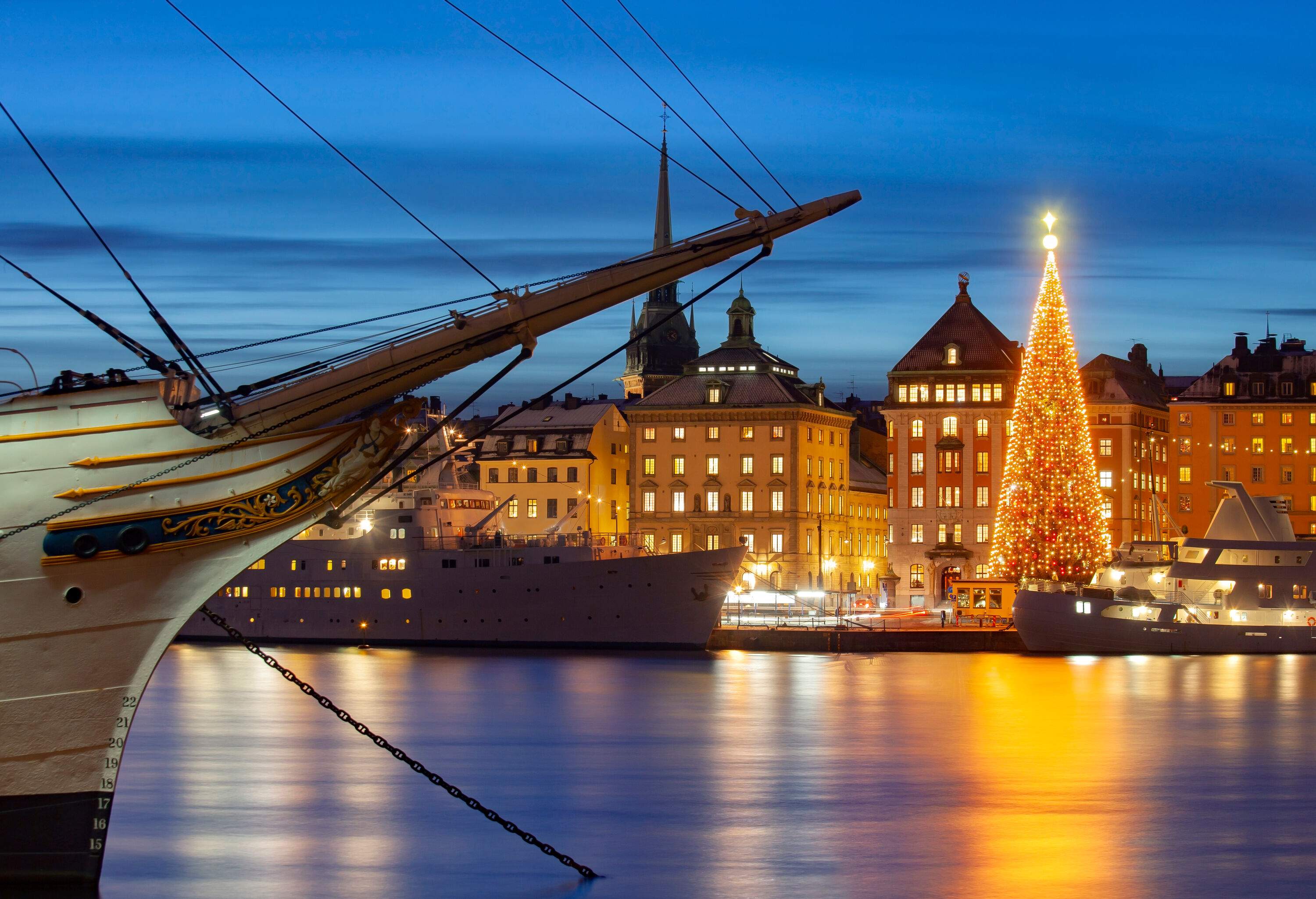 Dazzling Christmas tree and city buildings on a nearby pier with cruise ships and sailboats anchored on the harbour.