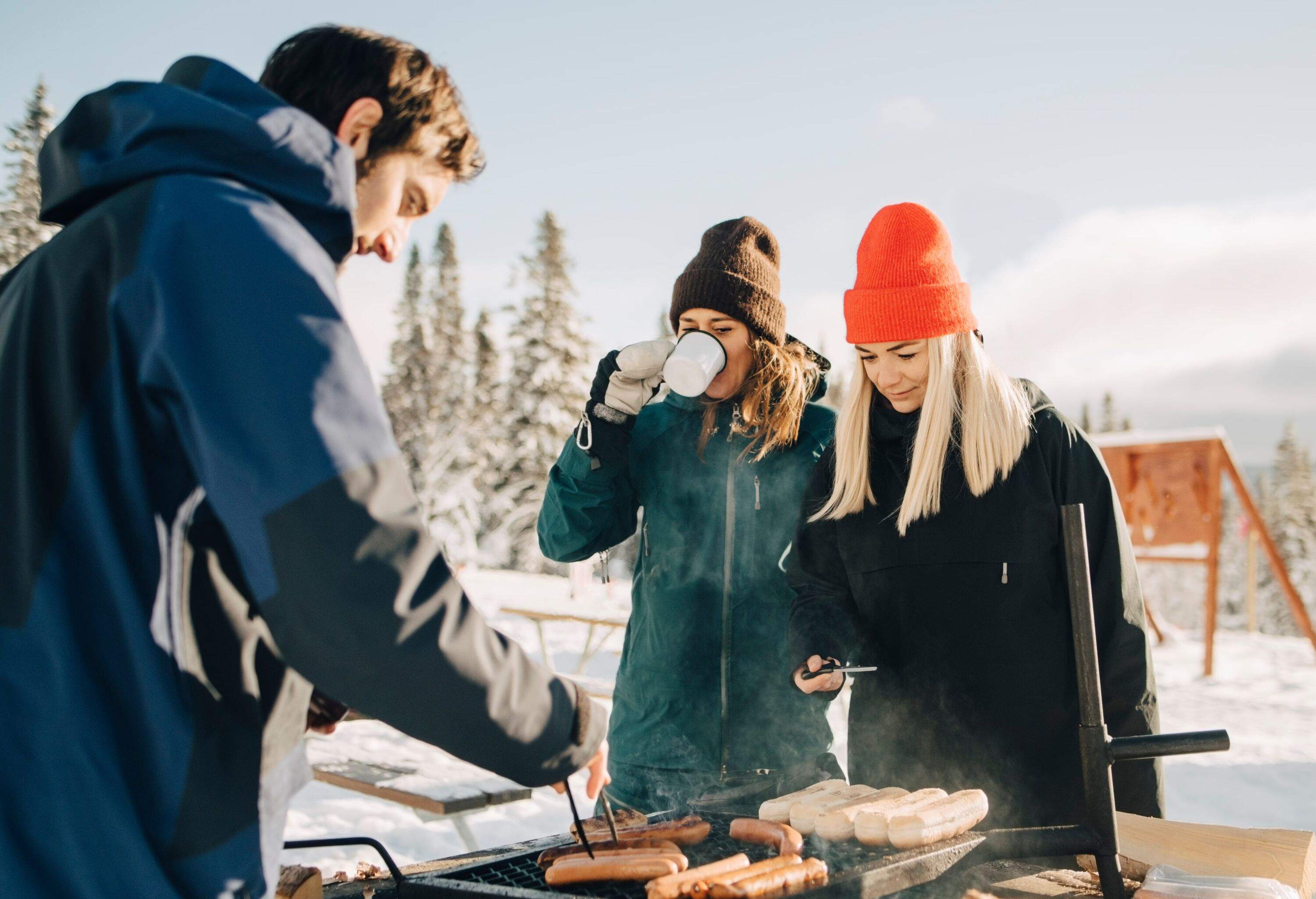 THREE FRIENDS IN SNOW GEAR ENJOYING AN OUTDOOR BARBEQUE ON A SUNNY WINTER SNOWY DAY