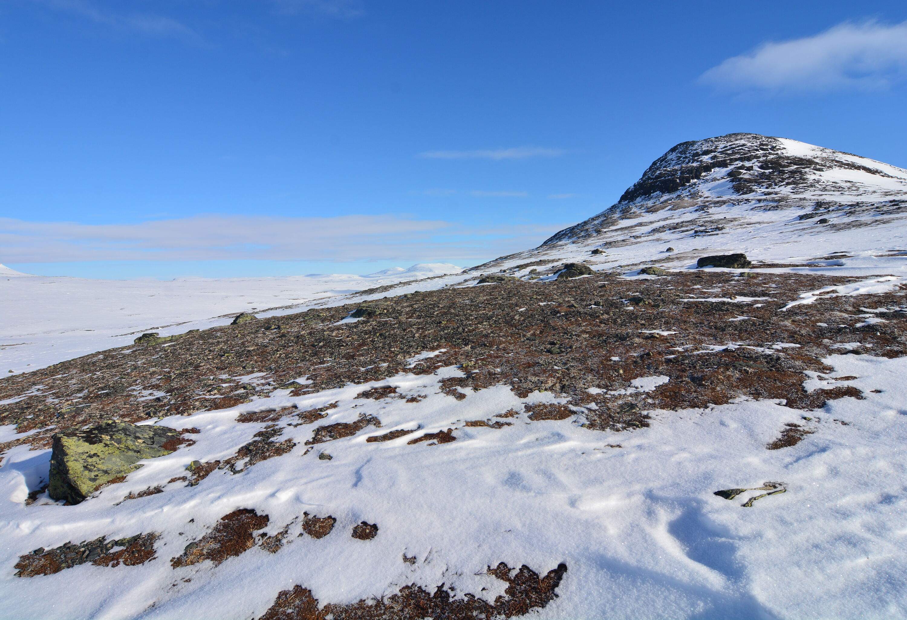 A sloping rocky hill partially covered with snow under the clear blue sky.