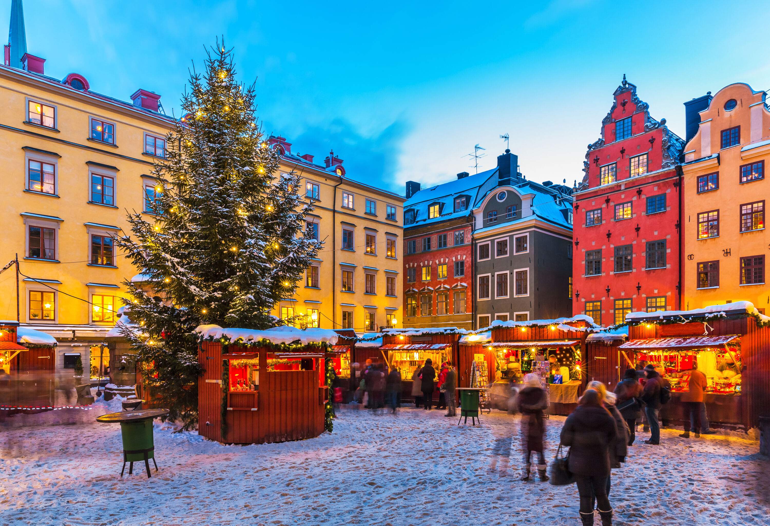 A brightly lit Christmas market with a tall Christmas tree in the centre of traditional iconic buildings covered in snow.