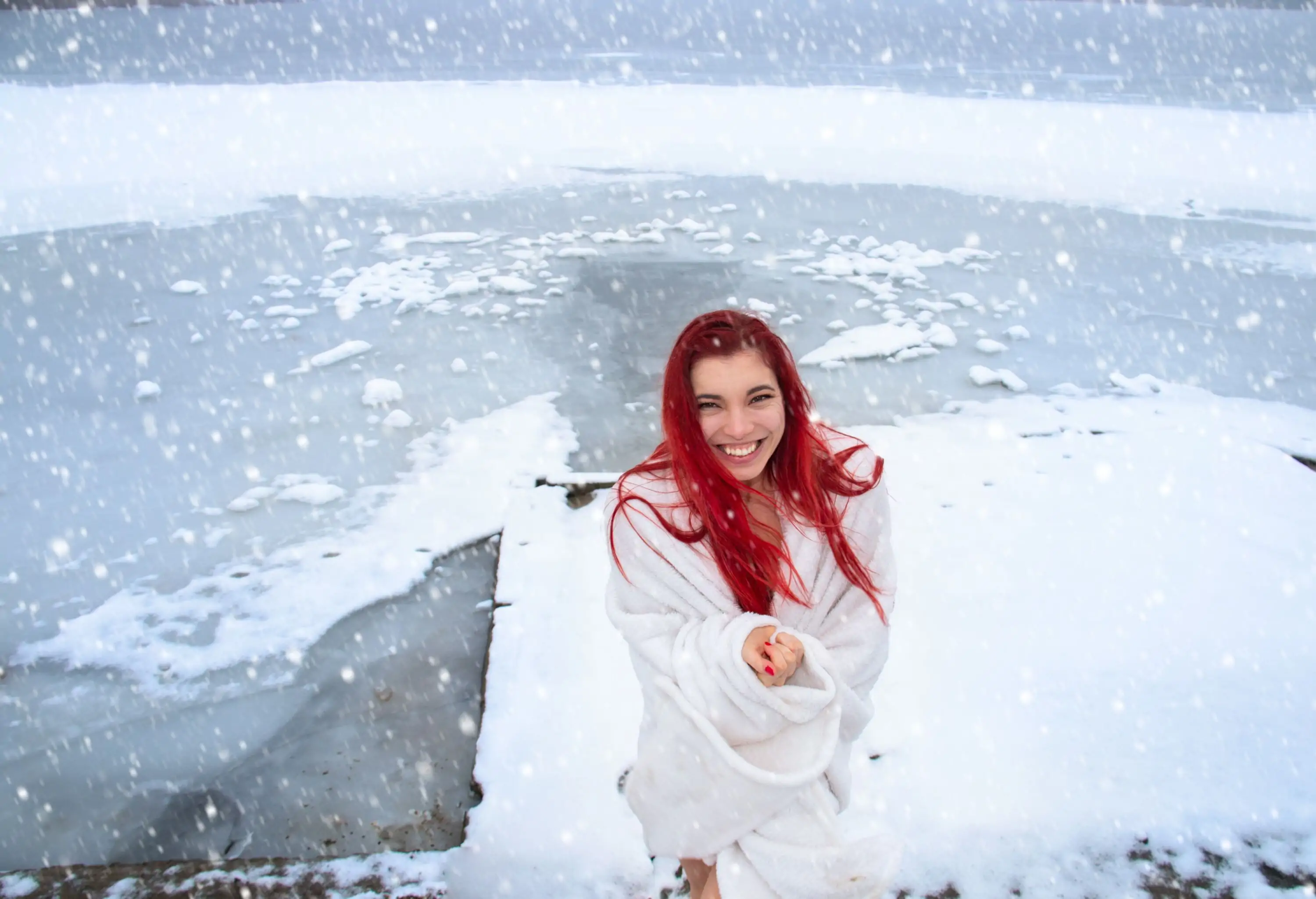 Happy young woman with long red hair, wrapped only in a blanket with a joyful smile enjoys natural cold therapy in winter outdoors and healthy lifestyle, hardening on snow, ice bath at the natural lake in snowy wild nature