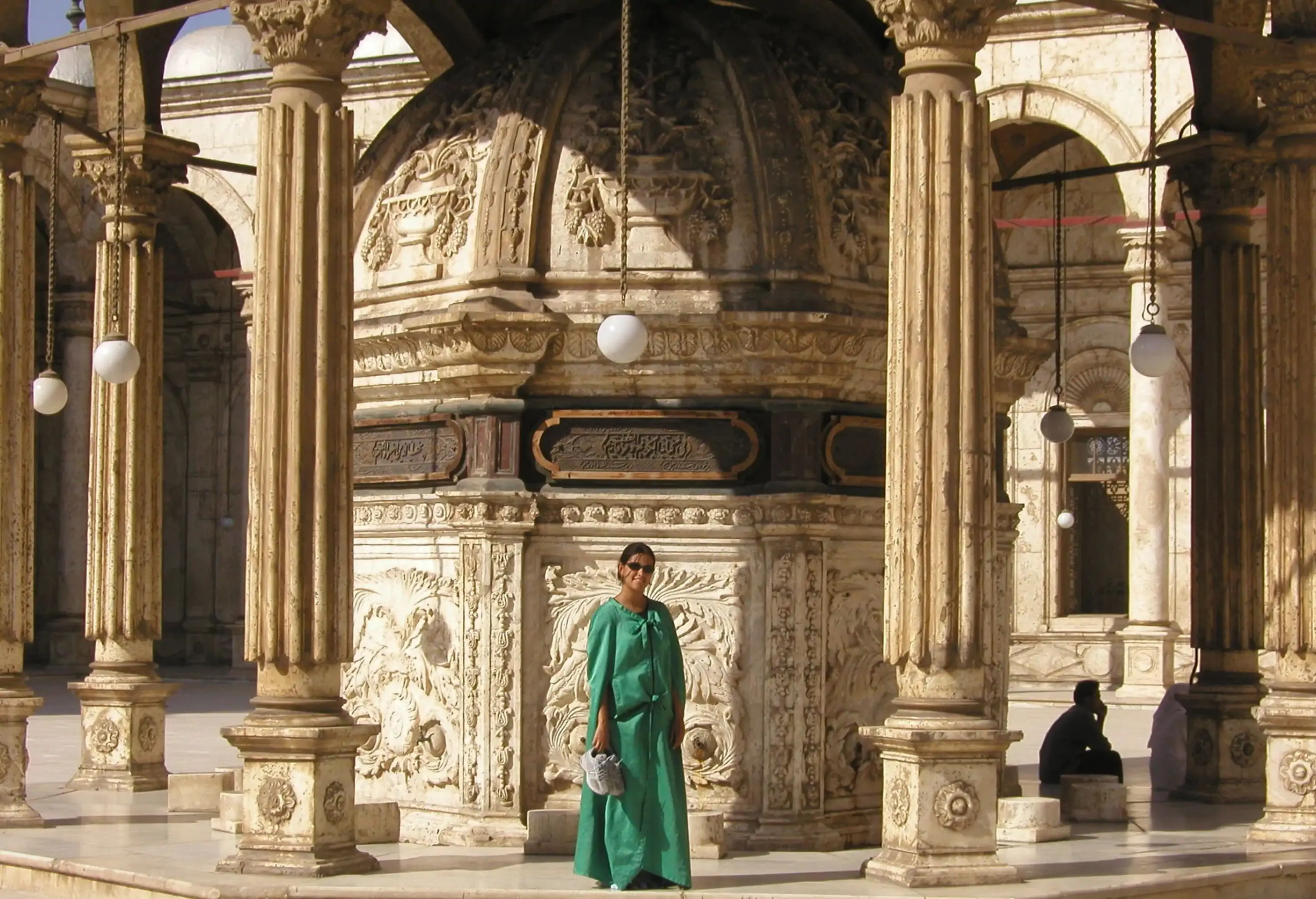 A lady tourist in a chic green dress poses against an ornate dome housed in a pavilion in the mosque courtyard.