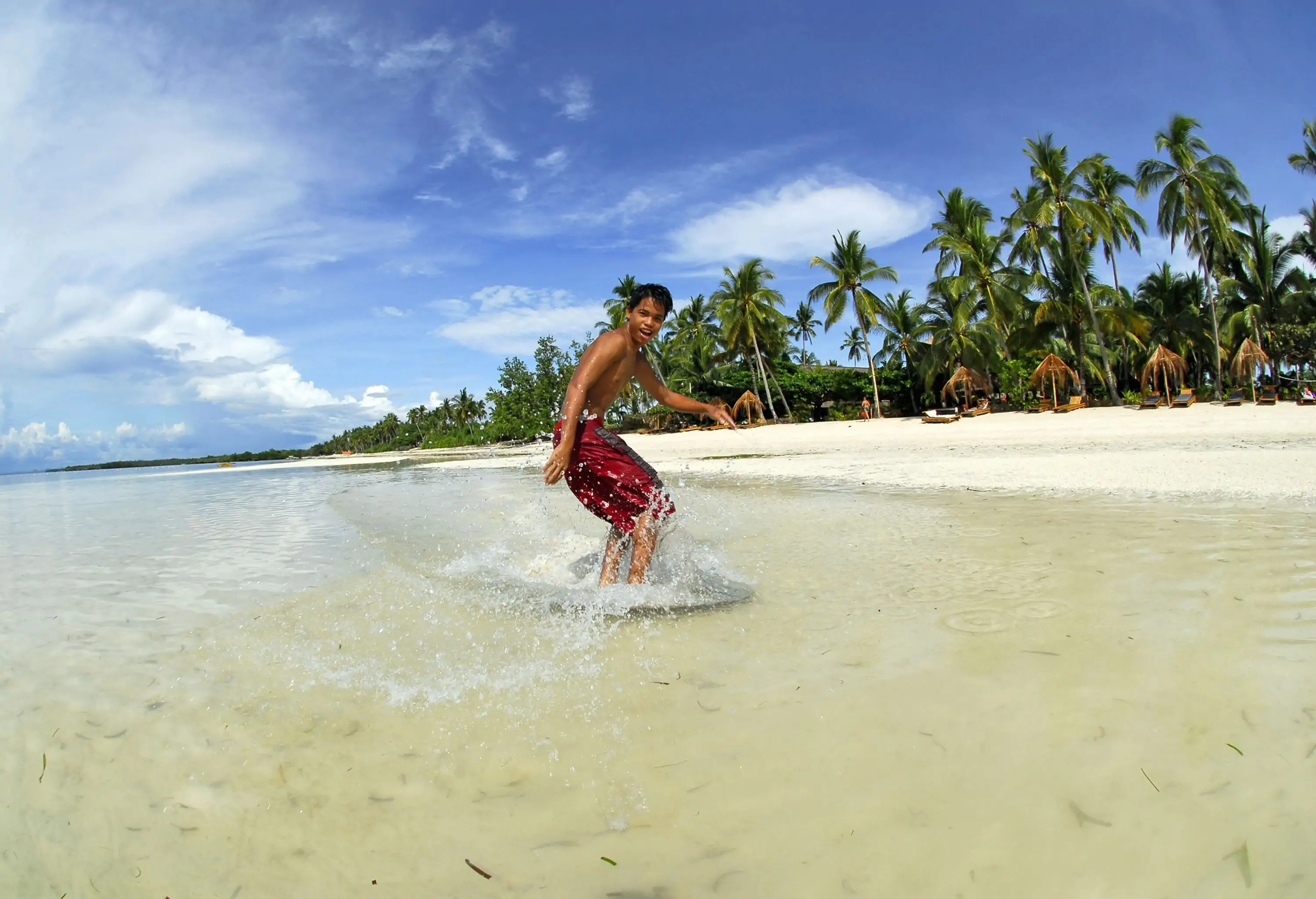 A young man wearing boardshorts skimboarding across the sea's surface near the shore.