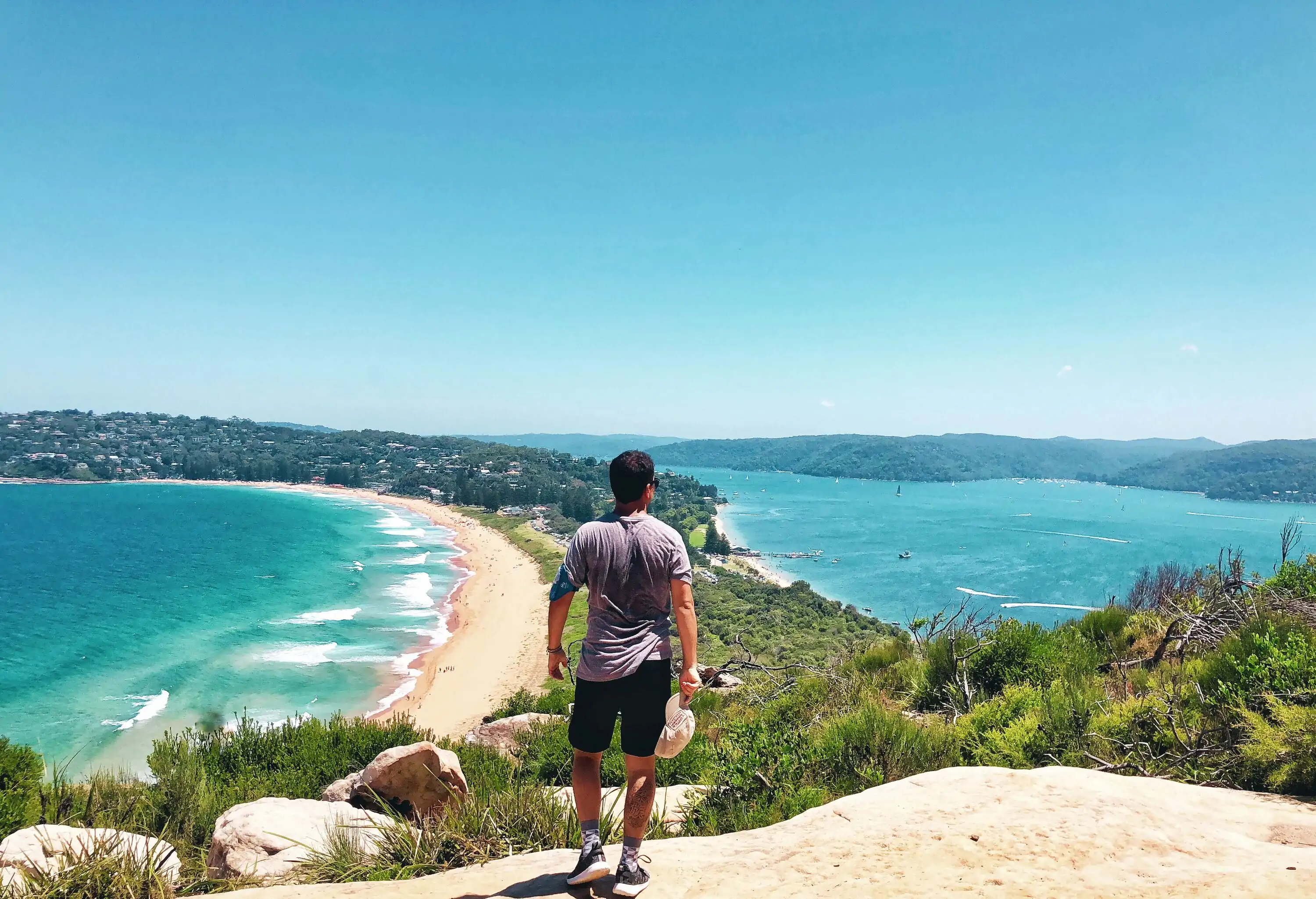 A man standing on top of a sand cliff overlooking a parallel beach with blue waters.