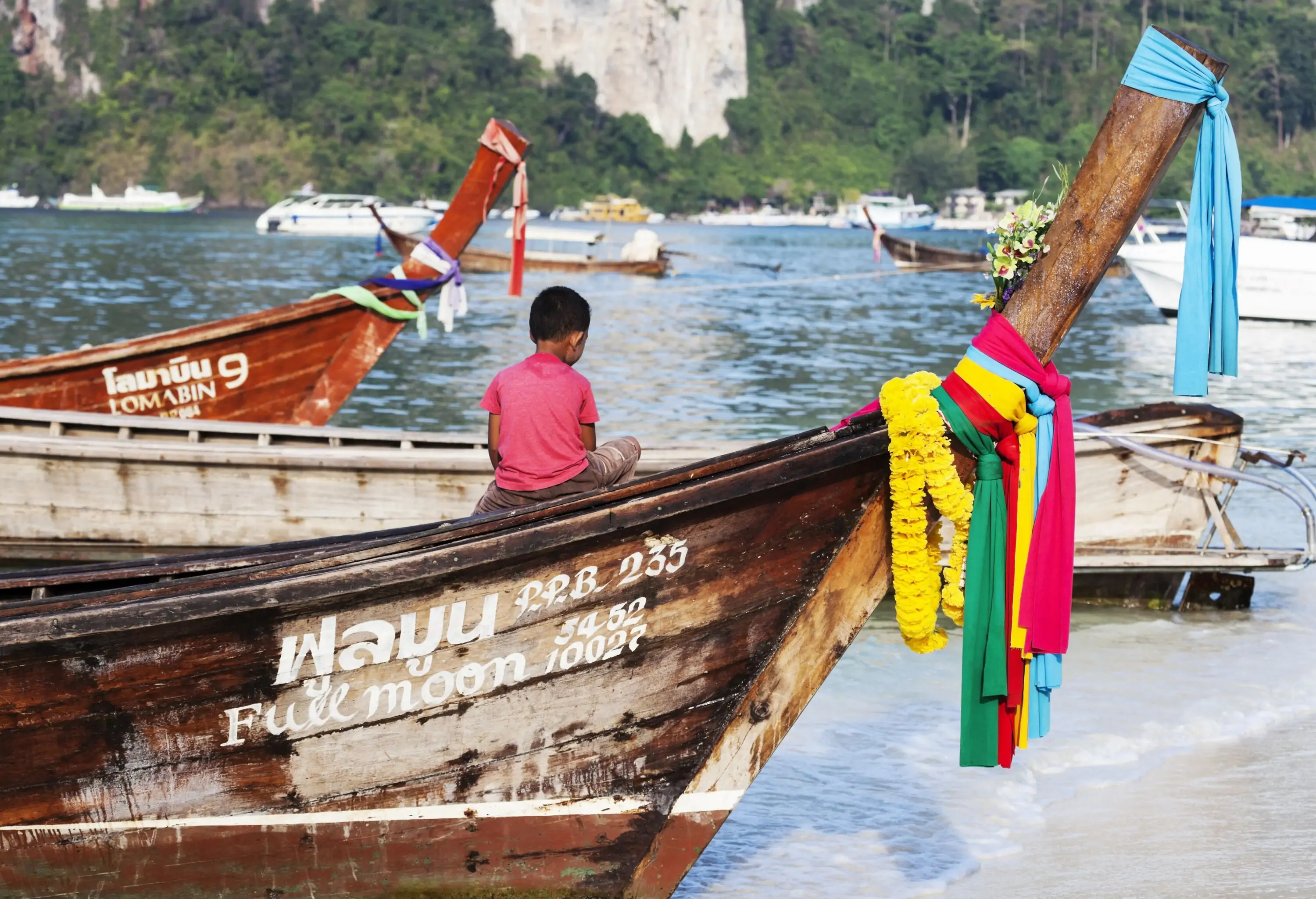 A little kid seat on a wooden boat resting on the shore of a beach.