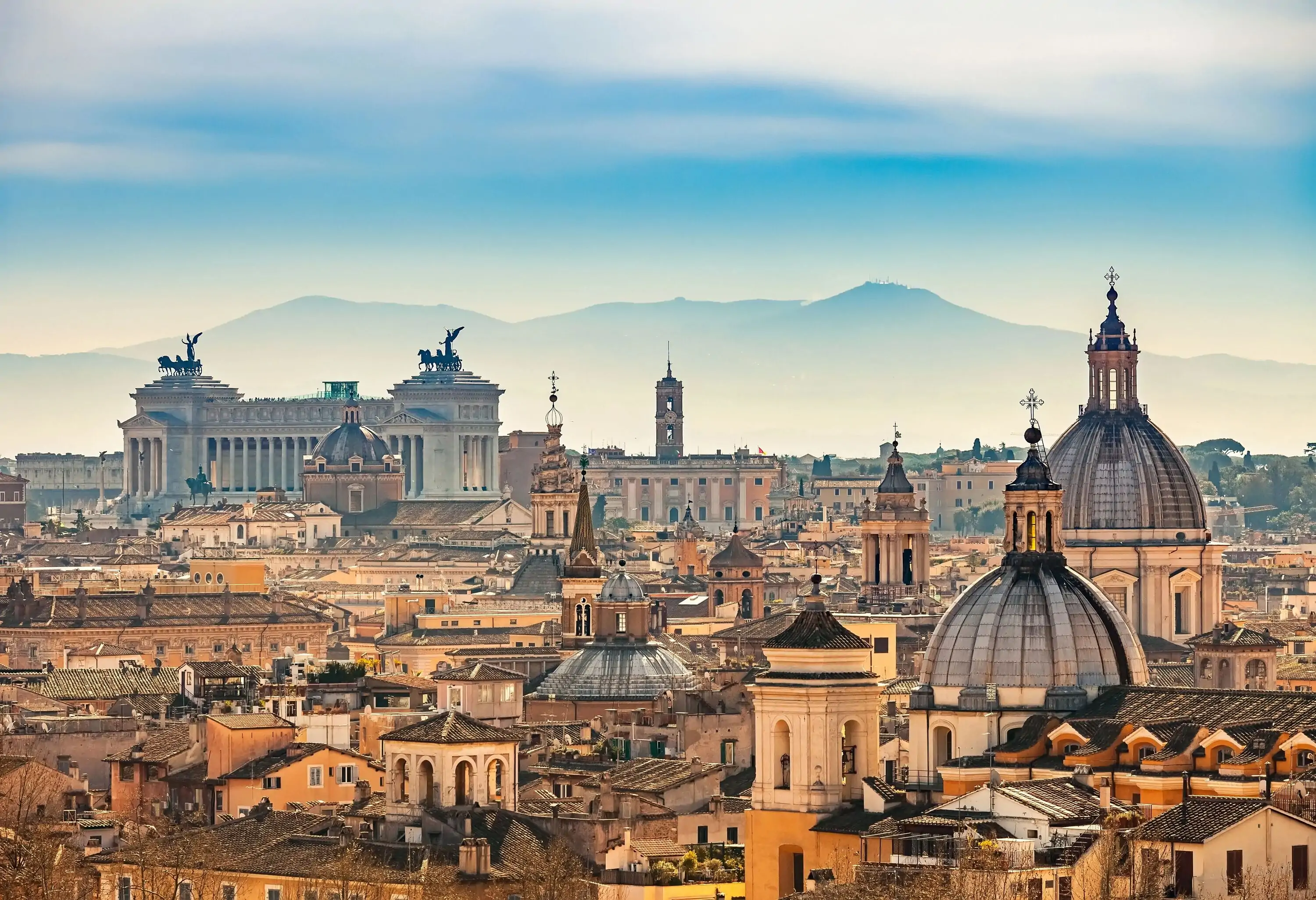 Amidst the bustling cityscape of Rome, two magnificent domed structures, along with the imposing Altar of the Fatherland, dominate the skyline, while in the distance, a serene and misty mountain range provides a stunning backdrop.