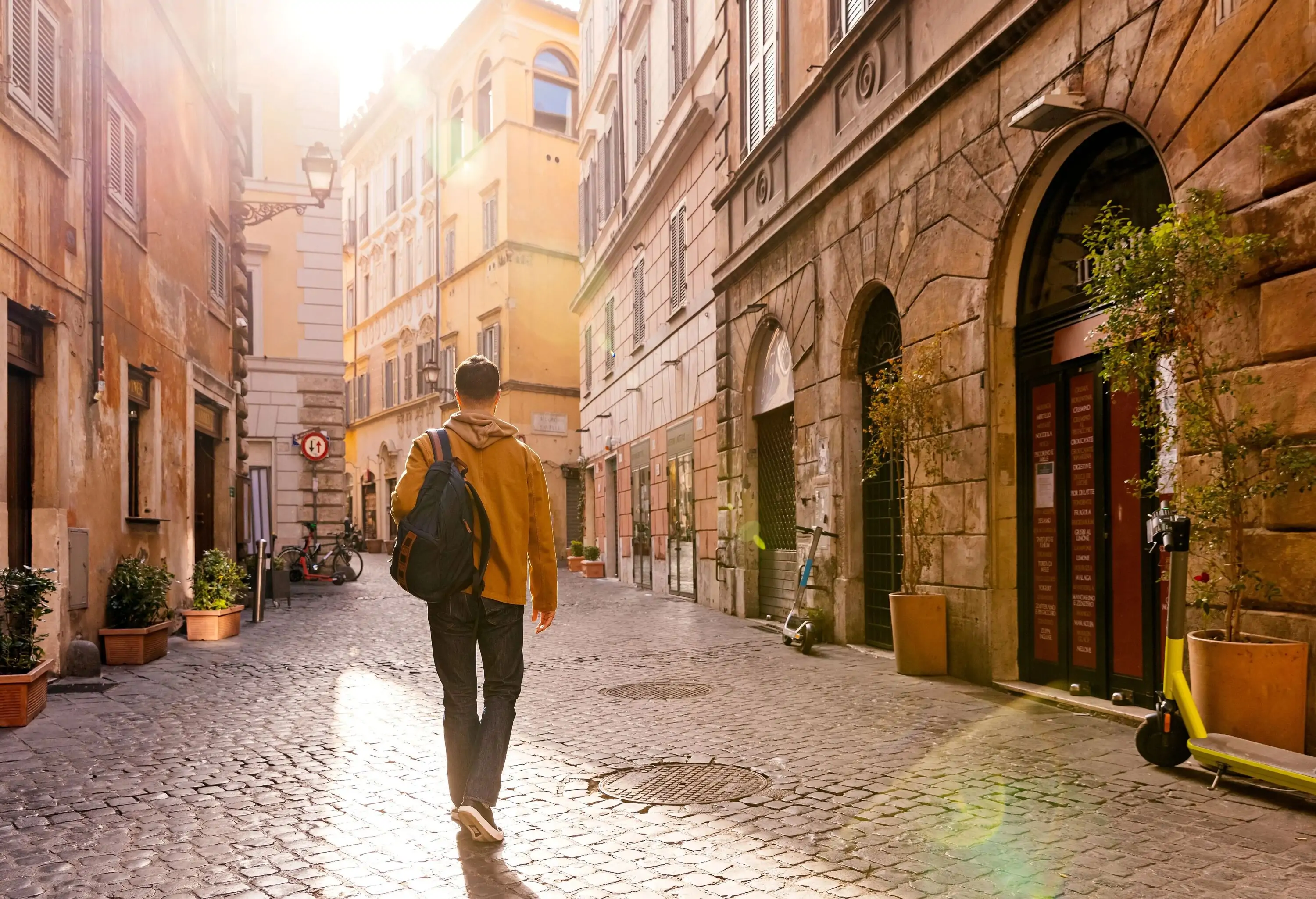 A man with a backpack walks on a sunlit cobbled street between tall buildings.