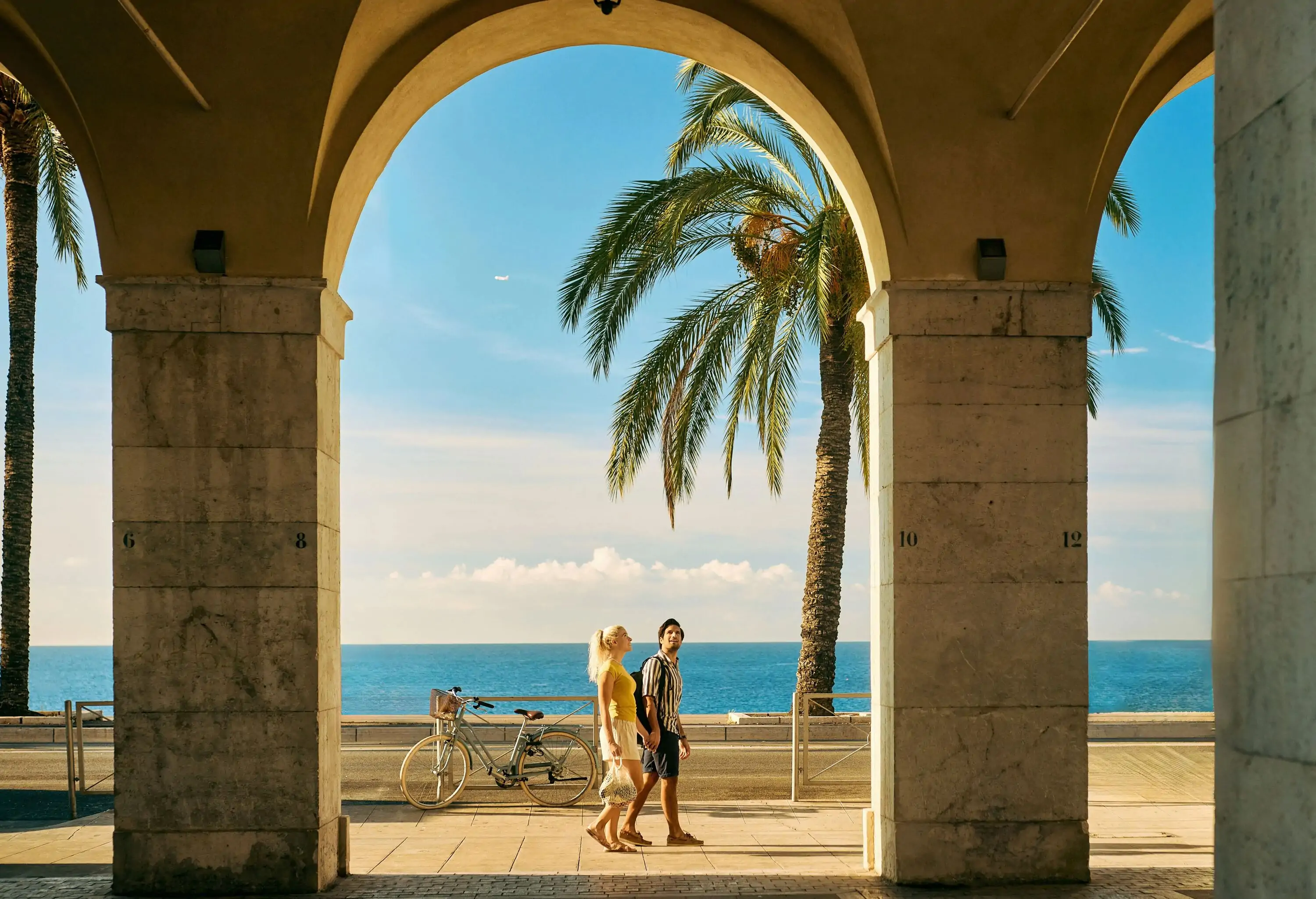 Wide shot of a couple on holiday walking under an archway on the Promenade des Anglais in Nice, France