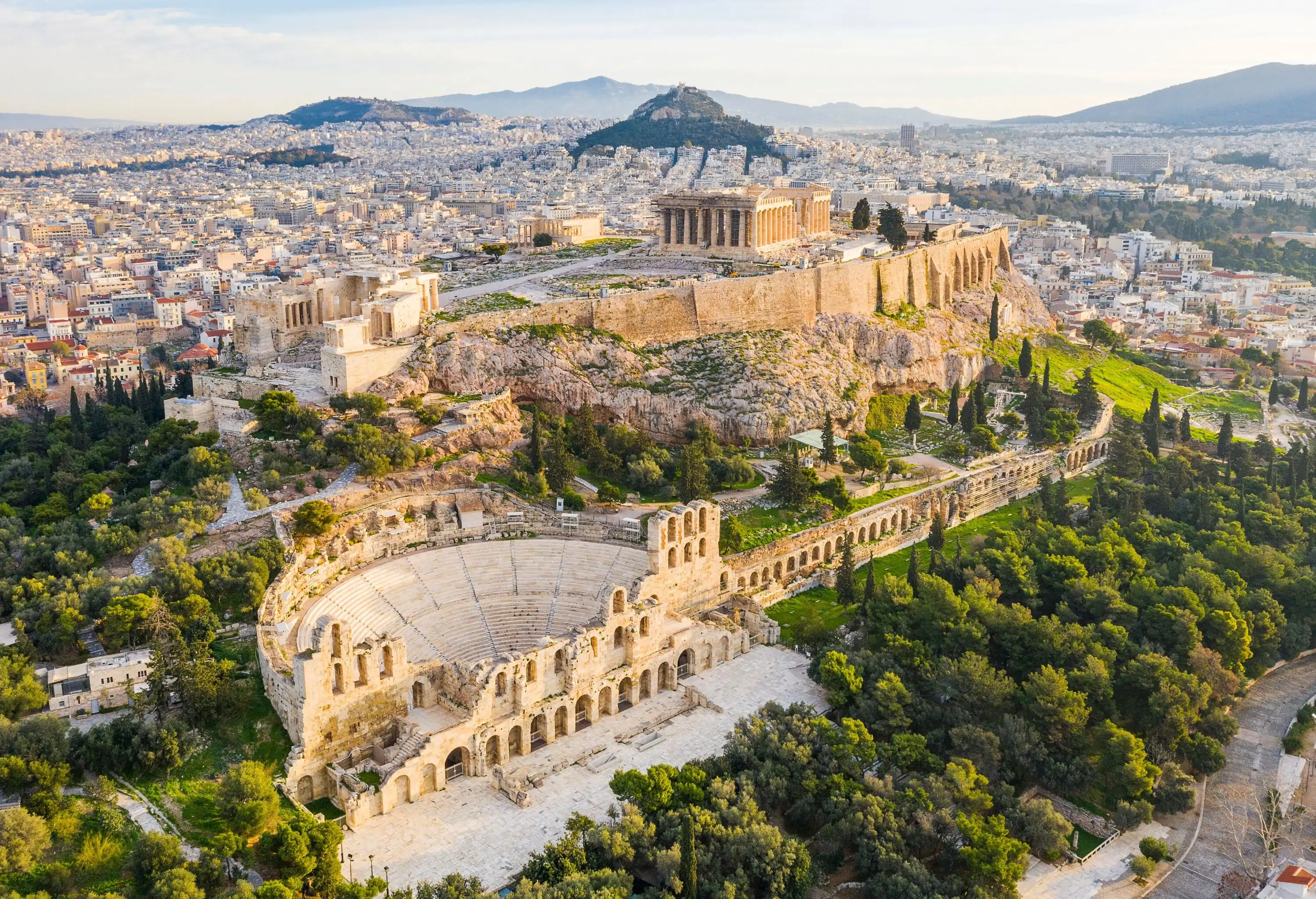 View of the city of Athens and the Acropolis in the centre above the Odeon of Herodes Atticus, a stone Roman theatre surrounded by lush tall trees.