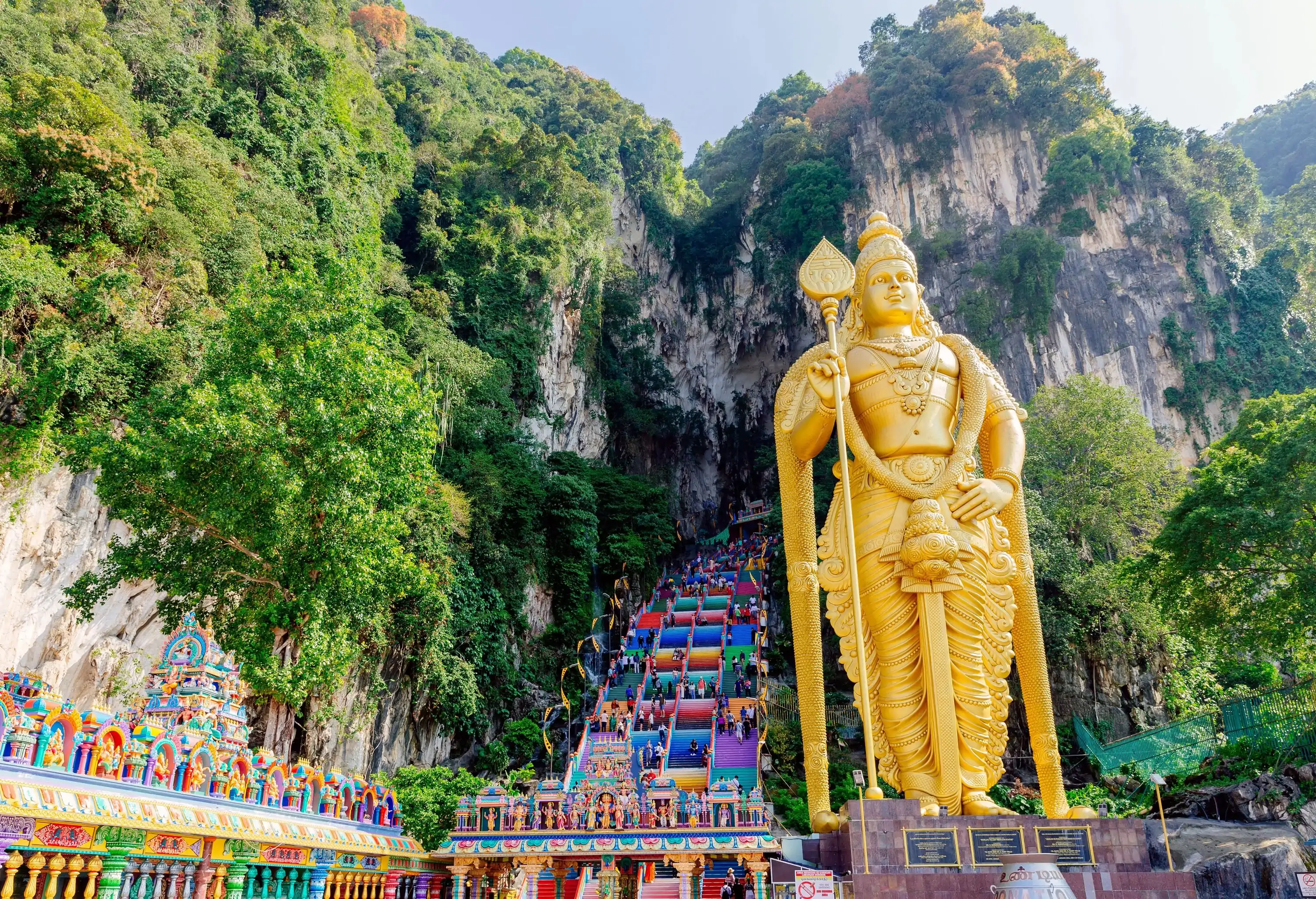 A monumental golden deity statue next to colourful and steep steps leading to a cave within a forest.